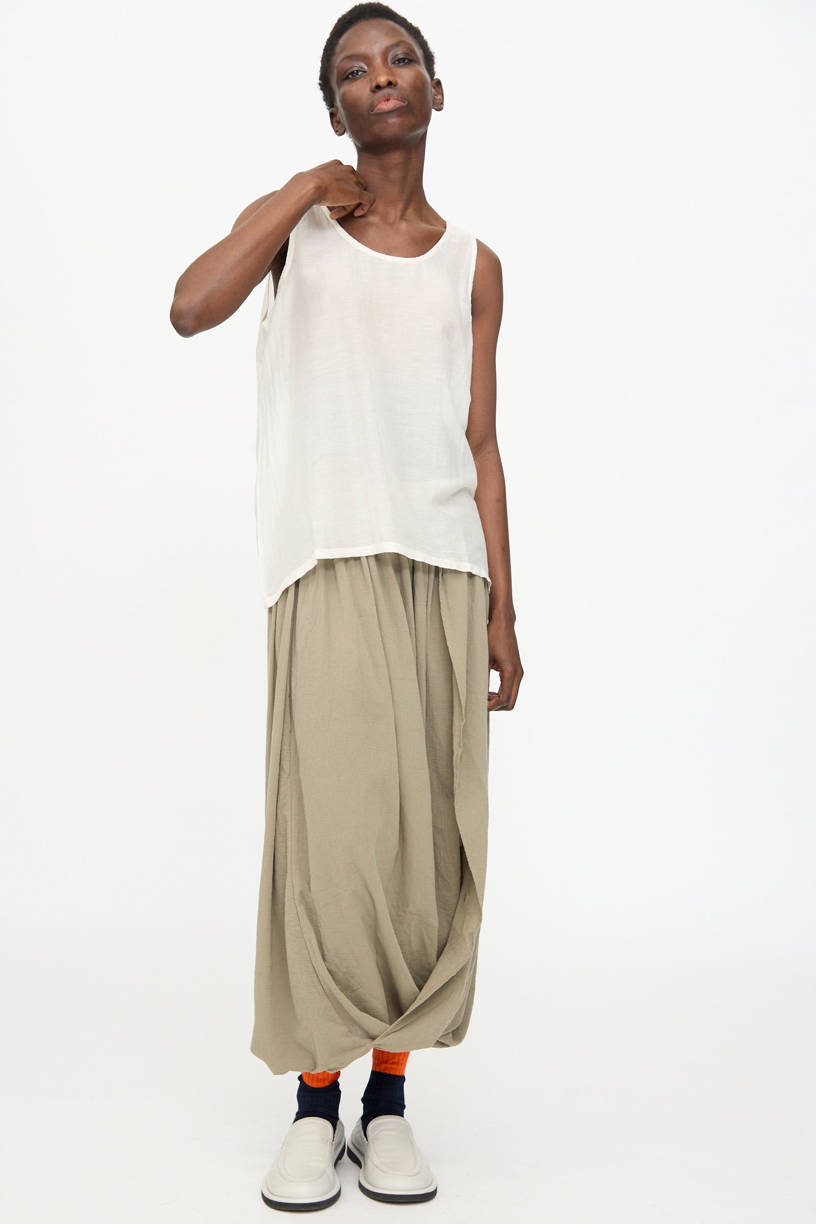 A person wears the Black Crane Loose Tank Top in White, beige draped pants, orange socks, and white slip-on shoes while standing against a plain white background.