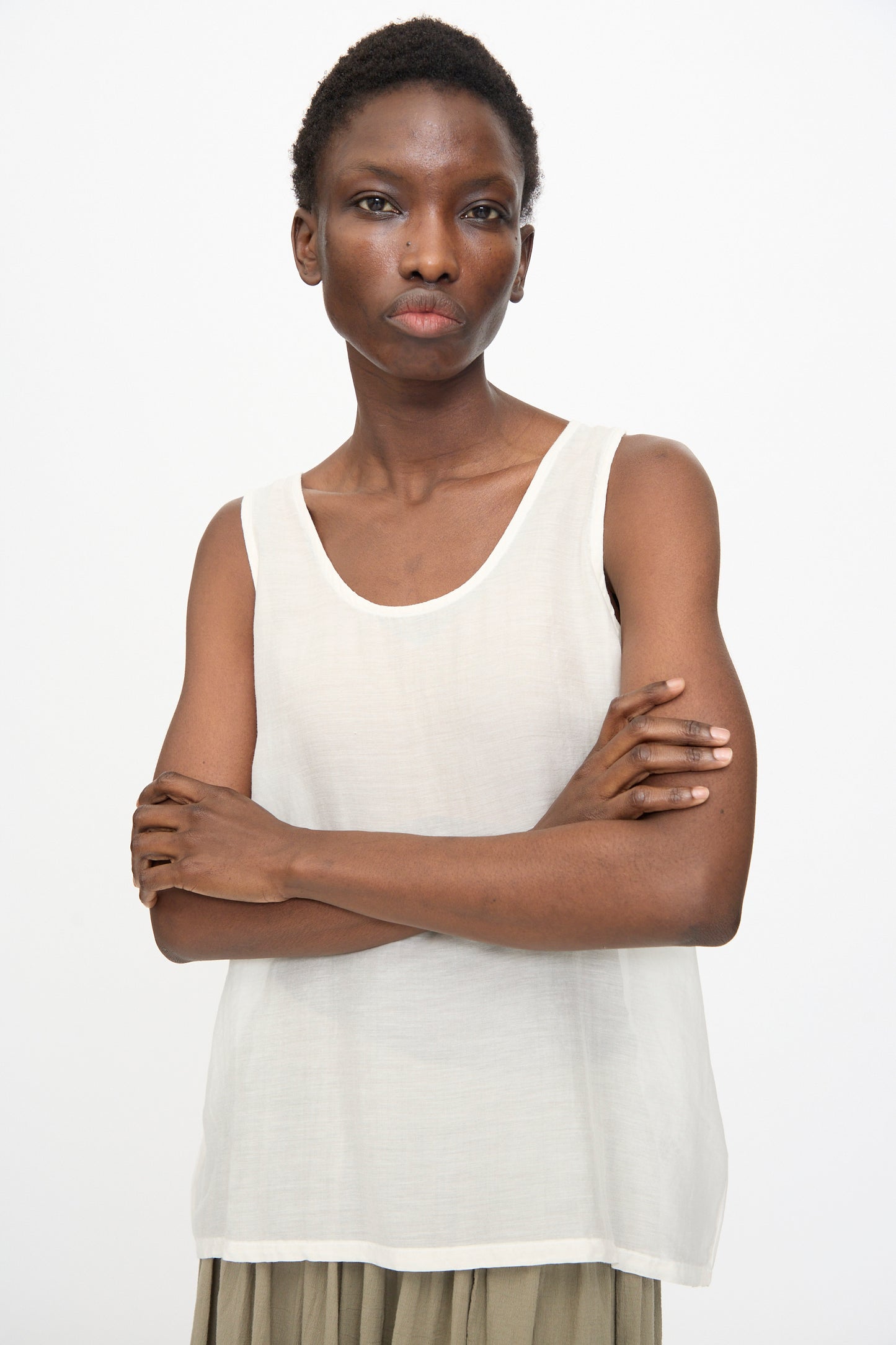 A person stands with arms crossed, wearing the Loose Tank Top in White by Black Crane and light-colored pants, looking directly at the camera against a plain background.