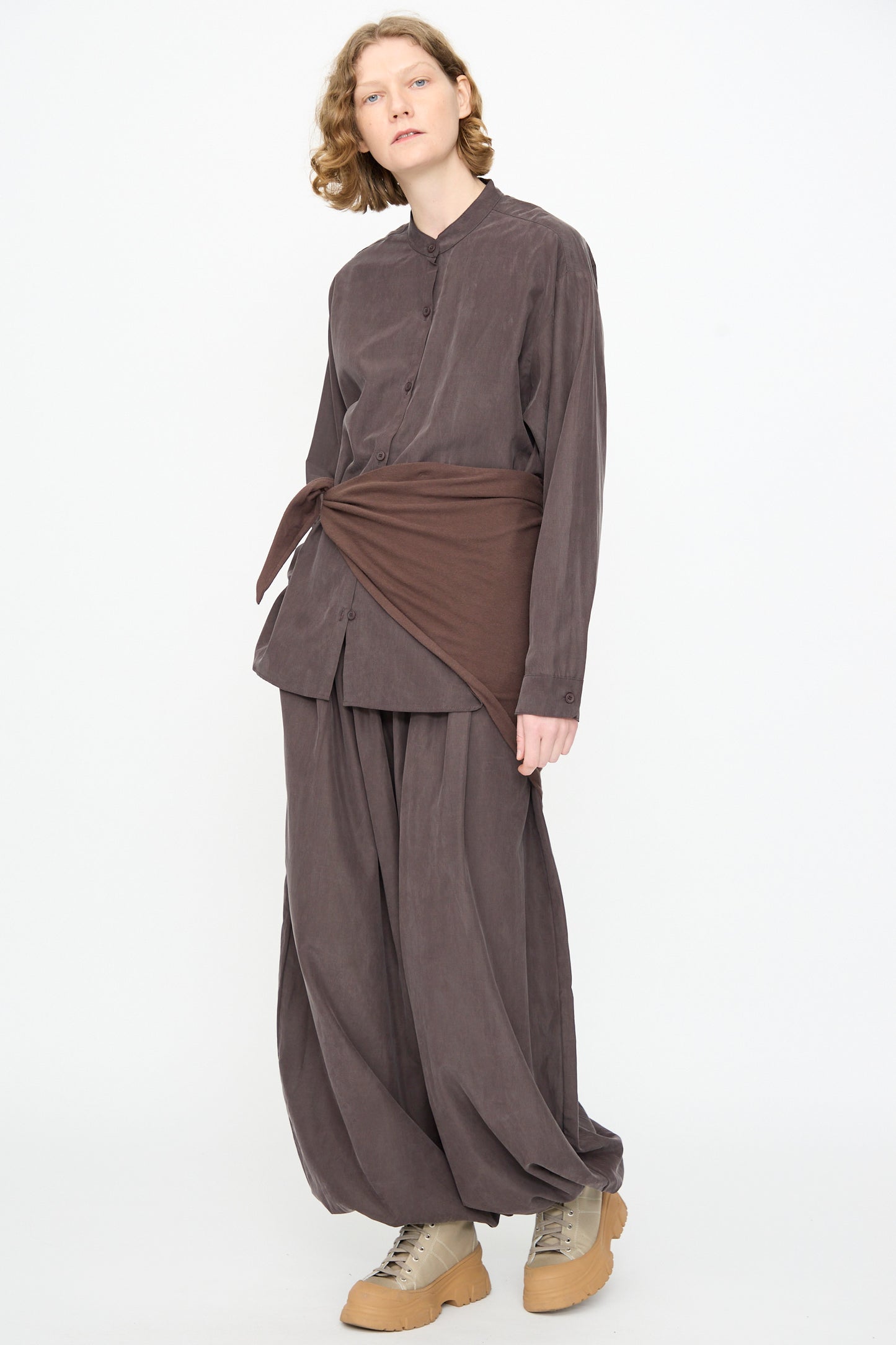 A person wearing the Black Crane Mao Collar Shirt in Dark Plum—a loose-fitting, band collar button-up—paired with matching pants, crafted from a soft cotton cupro blend, styled with a brown wrap tied at the waist and beige shoes, stands against a plain white background.