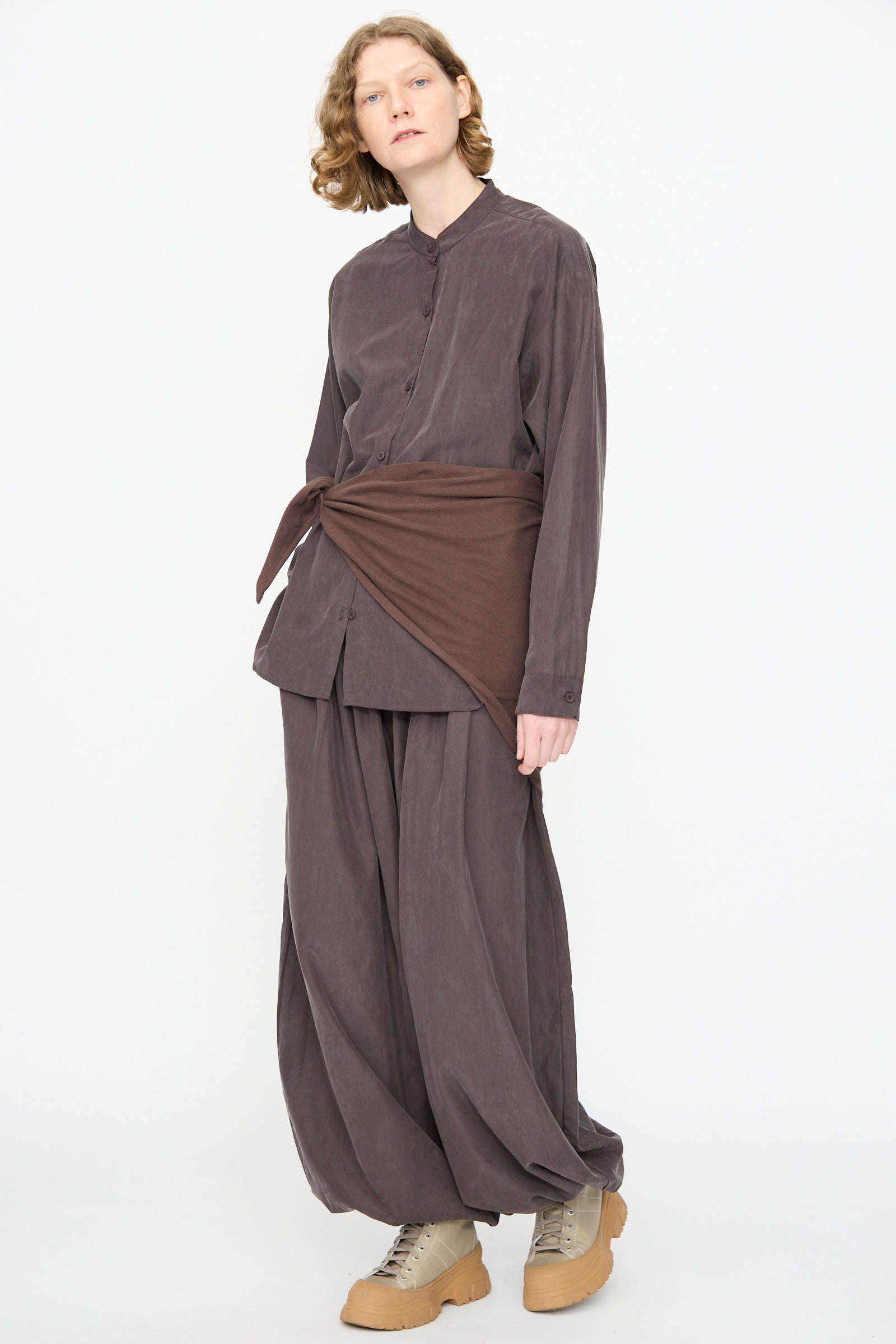A person wearing the Black Crane Mao Collar Shirt in Dark Plum—a loose-fitting, band collar button-up—paired with matching pants, crafted from a soft cotton cupro blend, styled with a brown wrap tied at the waist and beige shoes, stands against a plain white background.