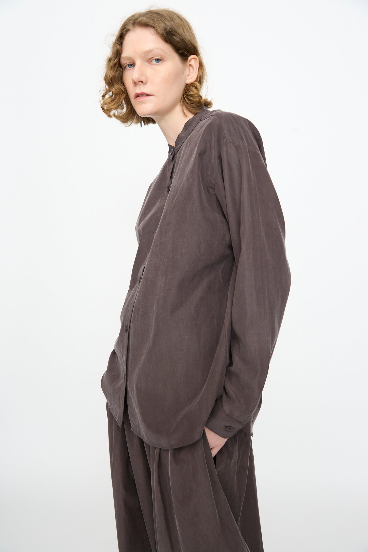 A person with wavy, shoulder-length hair is wearing the Black Crane Mao Collar Shirt in Dark Plum—a loose-fitting, long-sleeve button-up crafted from a soft cotton cupro blend—standing against a plain white background and looking slightly to the side.