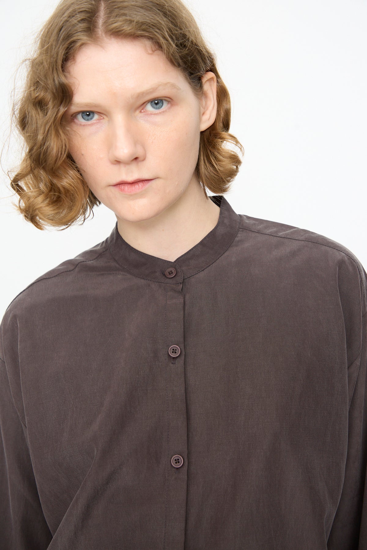A person with short, wavy blond hair is wearing the Black Crane Mao Collar Shirt in Dark Plum—a loose cotton cupro blend button-up with a band collar—while standing against a plain white background.