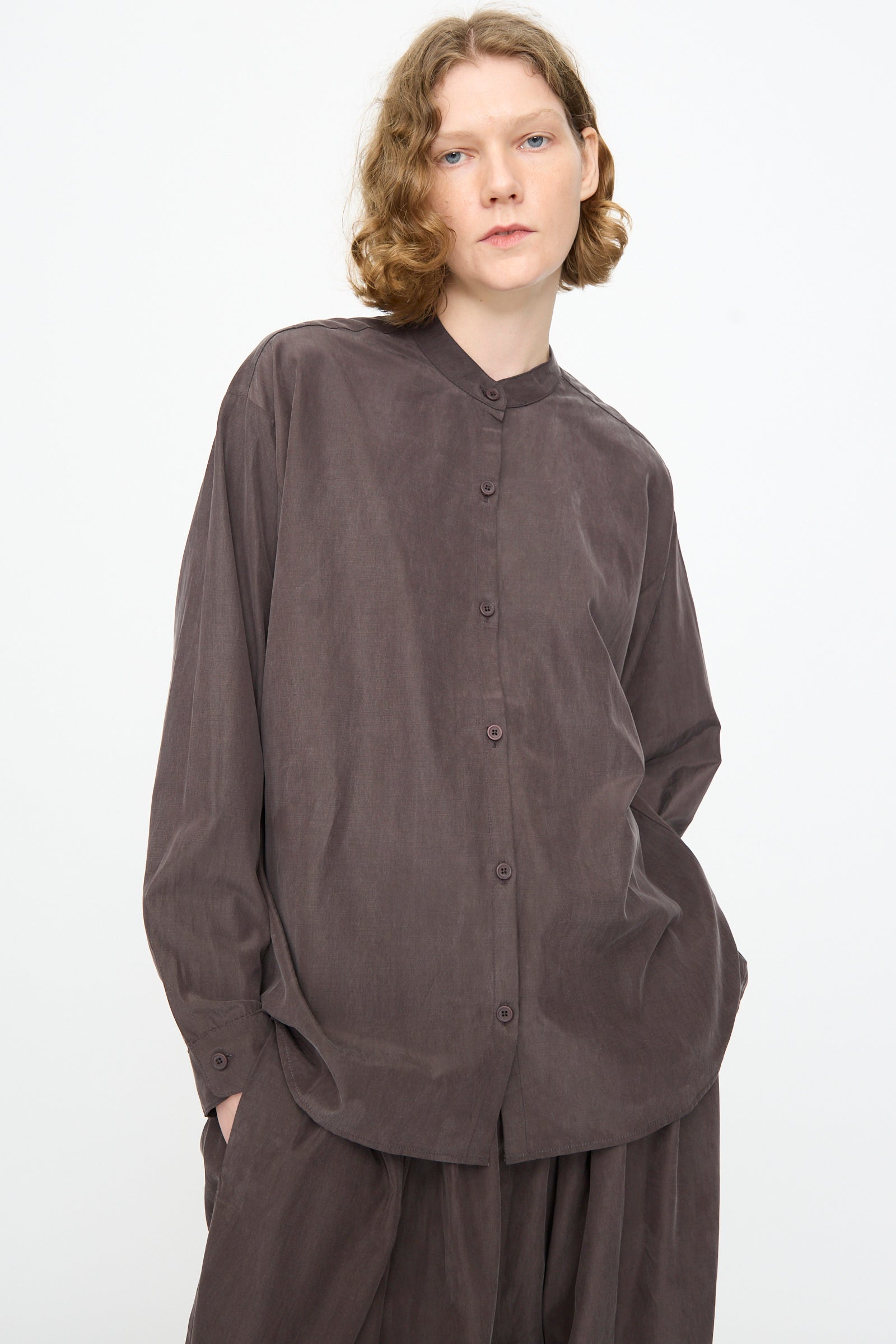 A person with shoulder-length wavy hair wears Black Crane’s Mao Collar Shirt in Dark Plum—a loose-fitting, button-up style crafted from a cotton cupro blend with a band collar—paired with matching dark brown pants, posing against a plain white background.