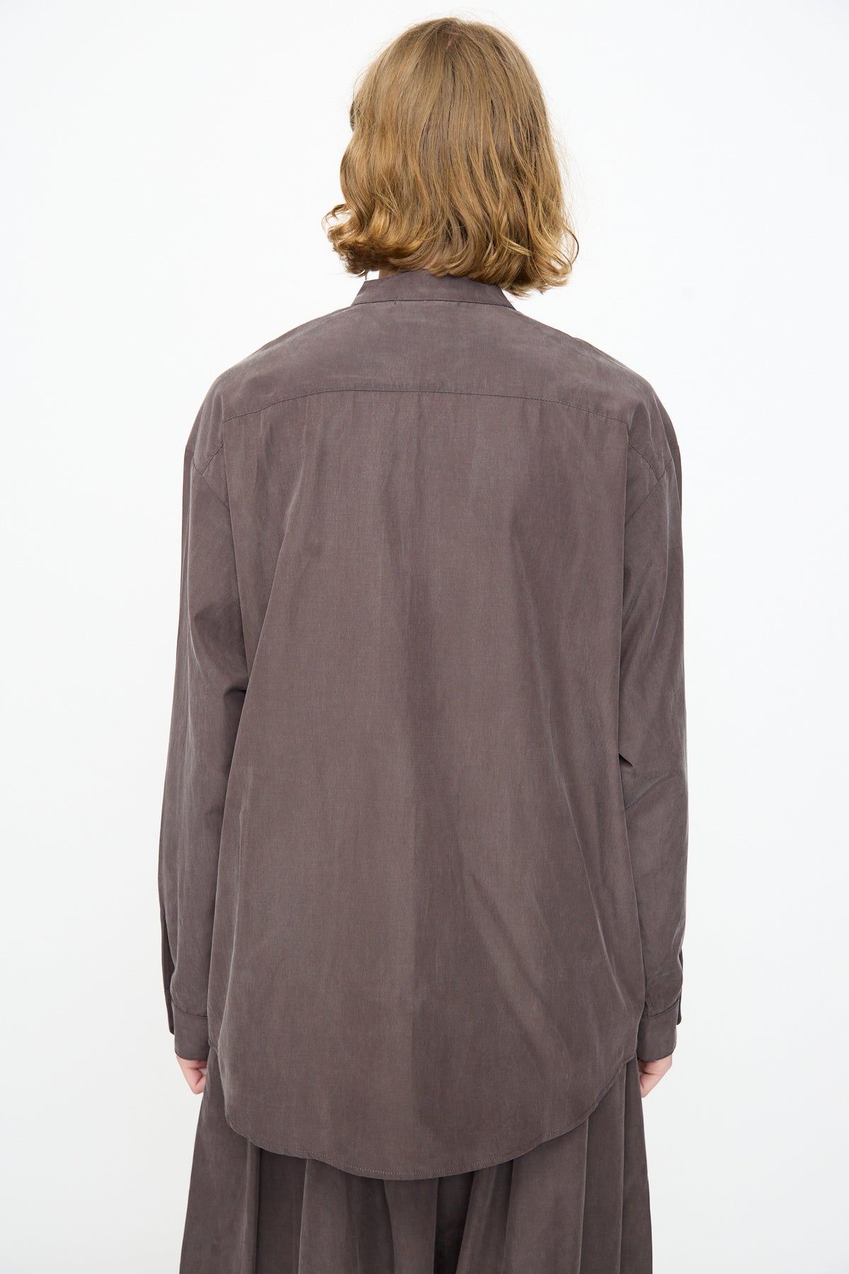 A person with shoulder-length light brown hair is seen from the back against a plain white background, wearing the Black Crane Mao Collar Shirt in Dark Plum—a loose-fitting, long-sleeve button-up crafted from a soft cotton cupro blend and featuring a band collar.