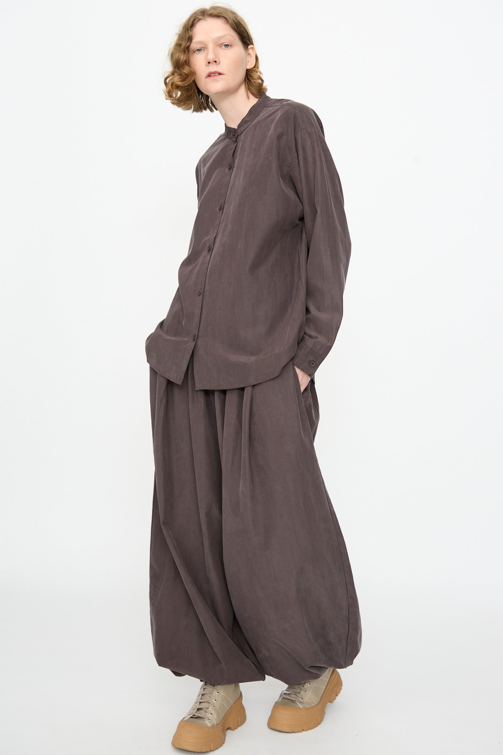 A person wearing the Black Crane Mao Collar Shirt in Dark Plum—a loose-fitting button-up with a band collar, crafted from a cotton cupro blend—paired with matching wide pants and tan shoes, stands with hands in pockets against a plain white background.