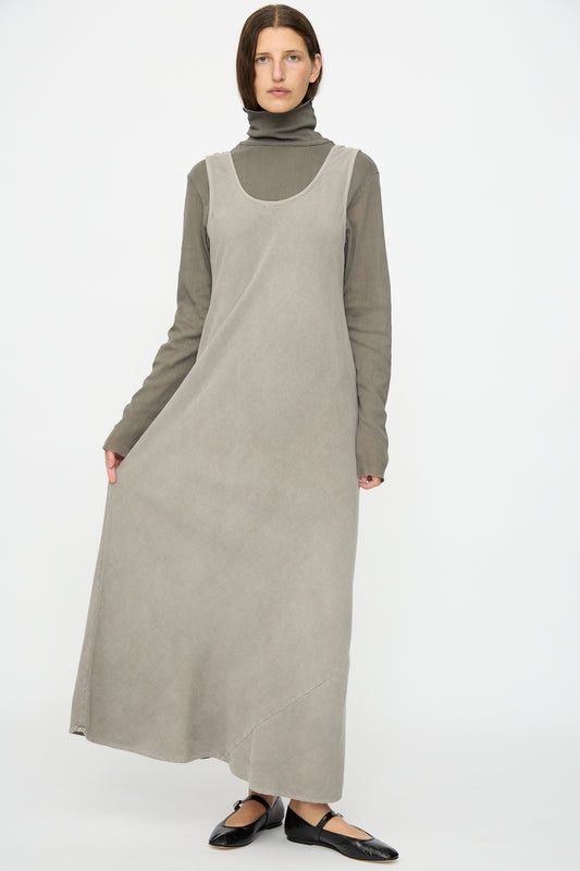 A person stands against a white background wearing the Black Crane Maxi Dress in Dark Stone, a relaxed fit, sleeveless style in gray cotton rayon blend, paired with a brown turtleneck top and black flat shoes.