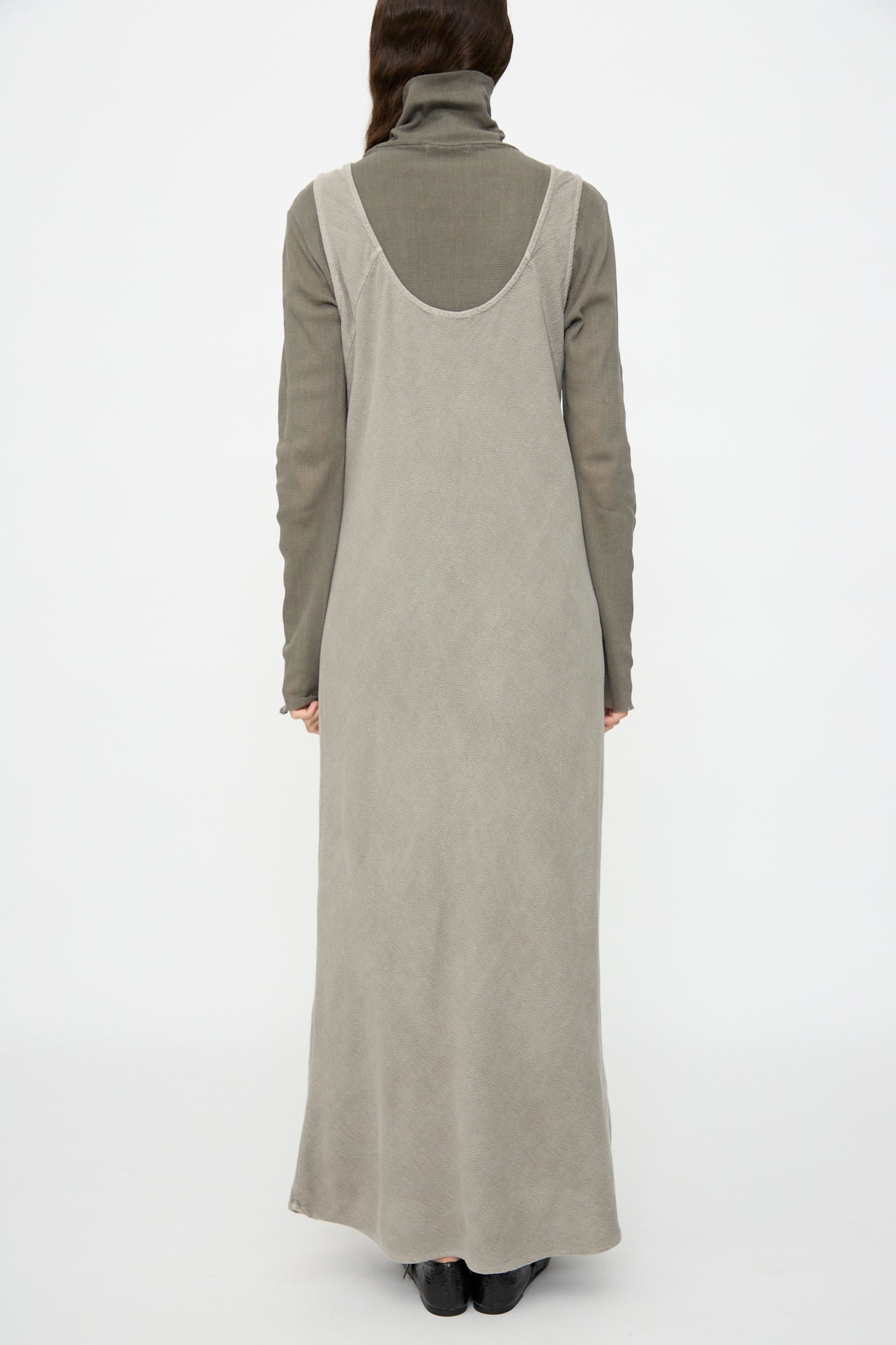 A person stands facing away, wearing the Black Crane Maxi Dress in Dark Stone—a relaxed fit, sleeveless maxi—layered over a gray long-sleeved turtleneck with black shoes, set against a plain white background.