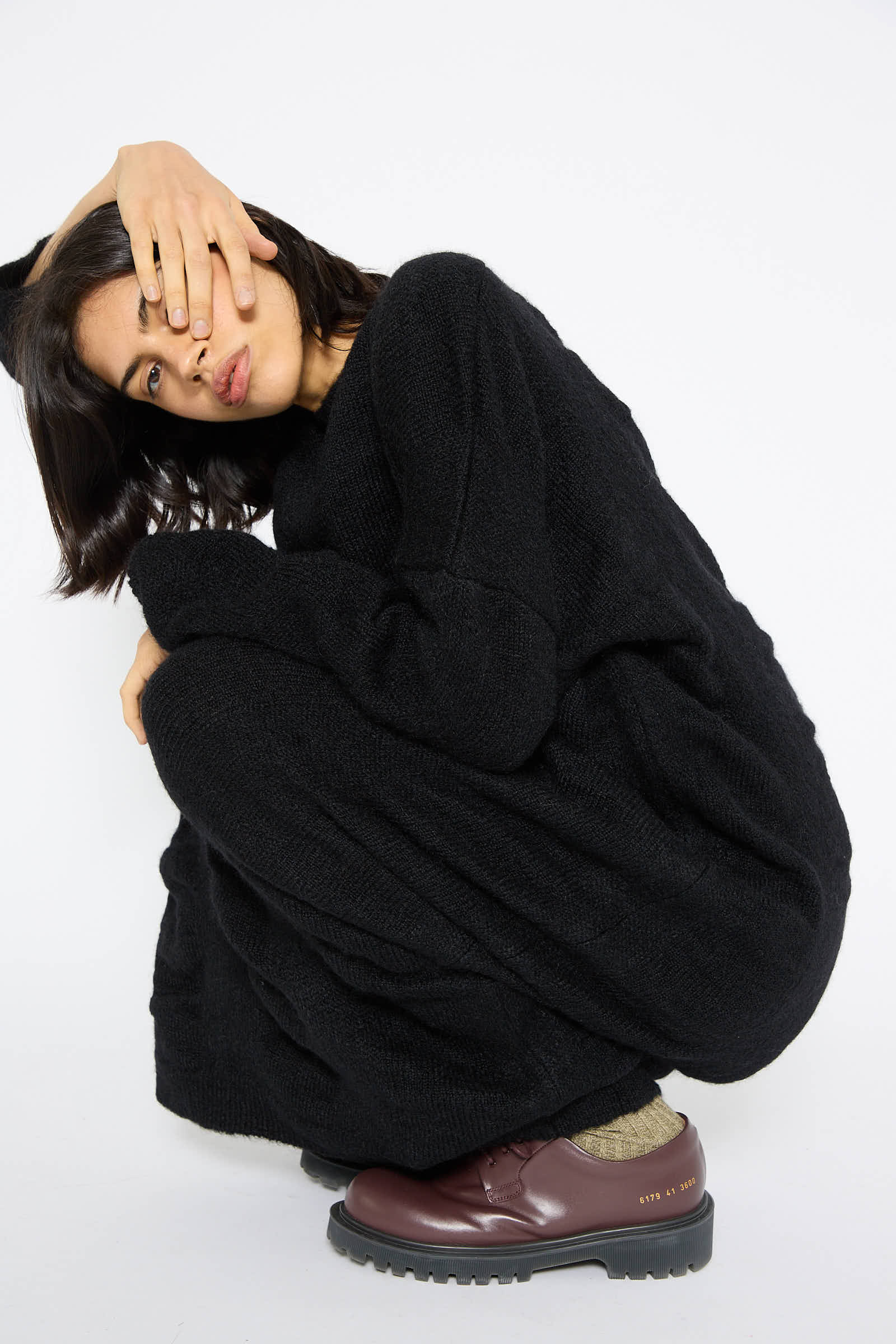 A person wearing a Black Crane Mohair and Wool Waterfall Knit Dress in black and brown shoes squats while covering one eye with their hand, exuding the elegance akin to a Japanese mohair blend, against a plain white background.