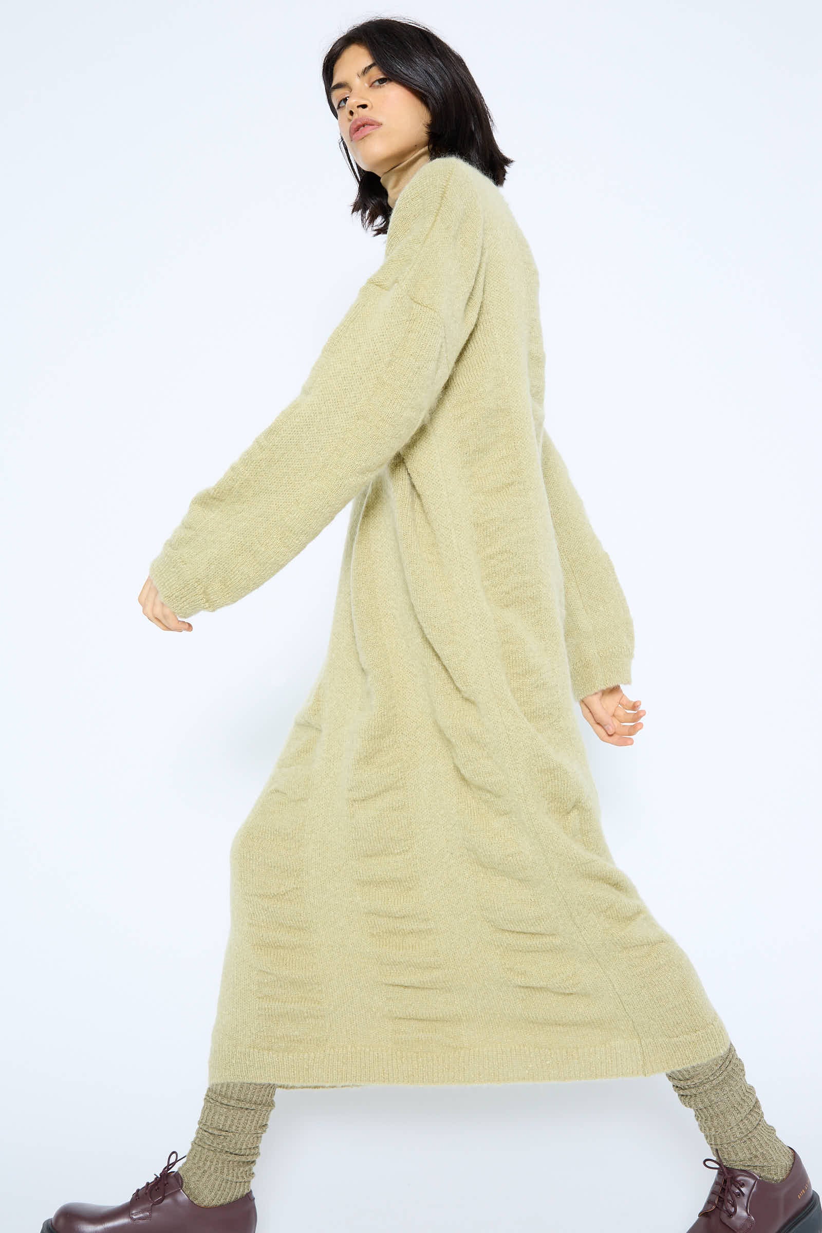 A person wearing a long, lime-colored Mohair and Wool Waterfall Knit Dress by Black Crane, brown lace-up boots, and beige knitted leg warmers is walking, shown in a side profile against a light background.