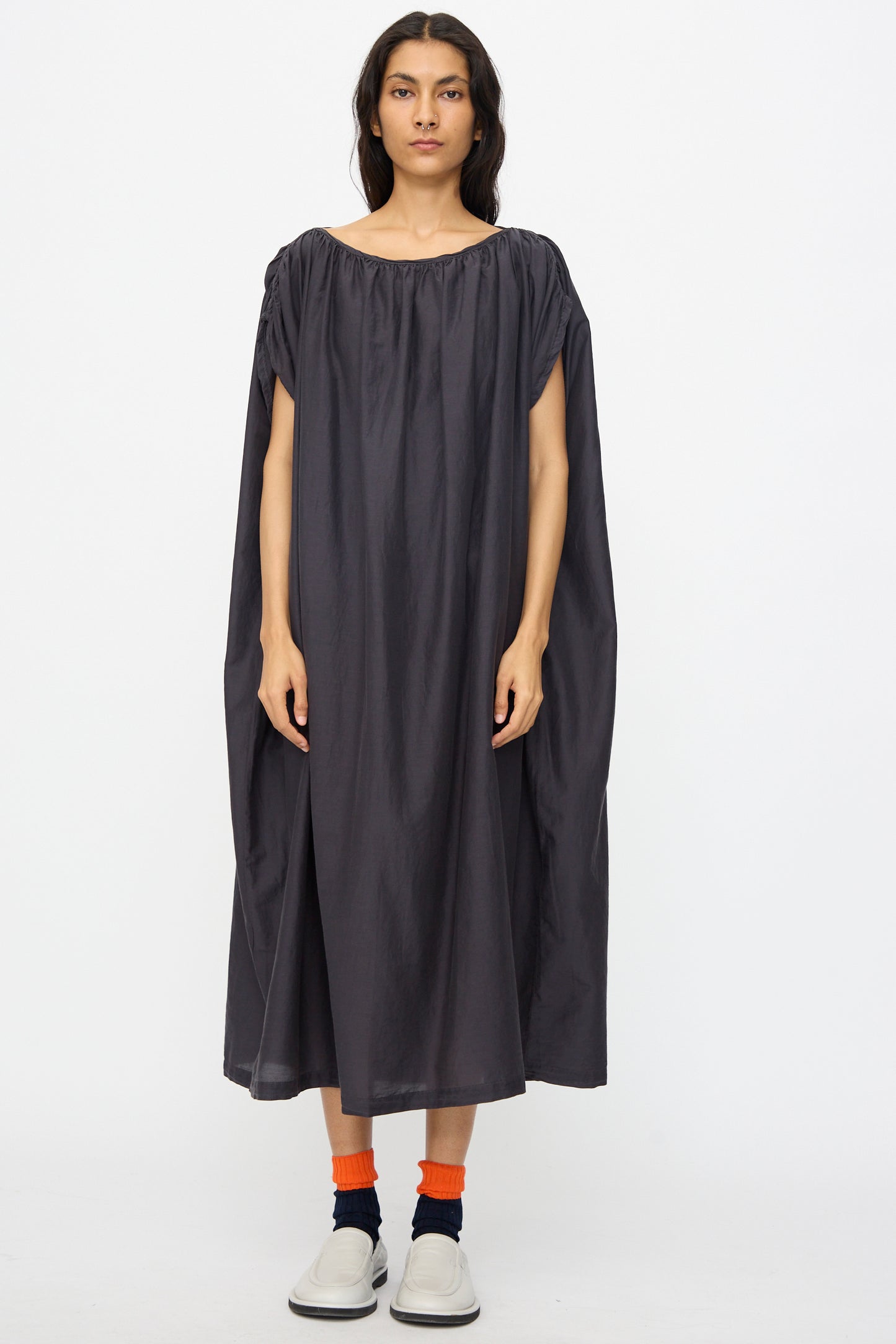 Person stands against a white background wearing the Black Crane Mont Blanc Dress in Faded Black, featuring an oversized silhouette, paired with orange socks and white slip-on shoes.