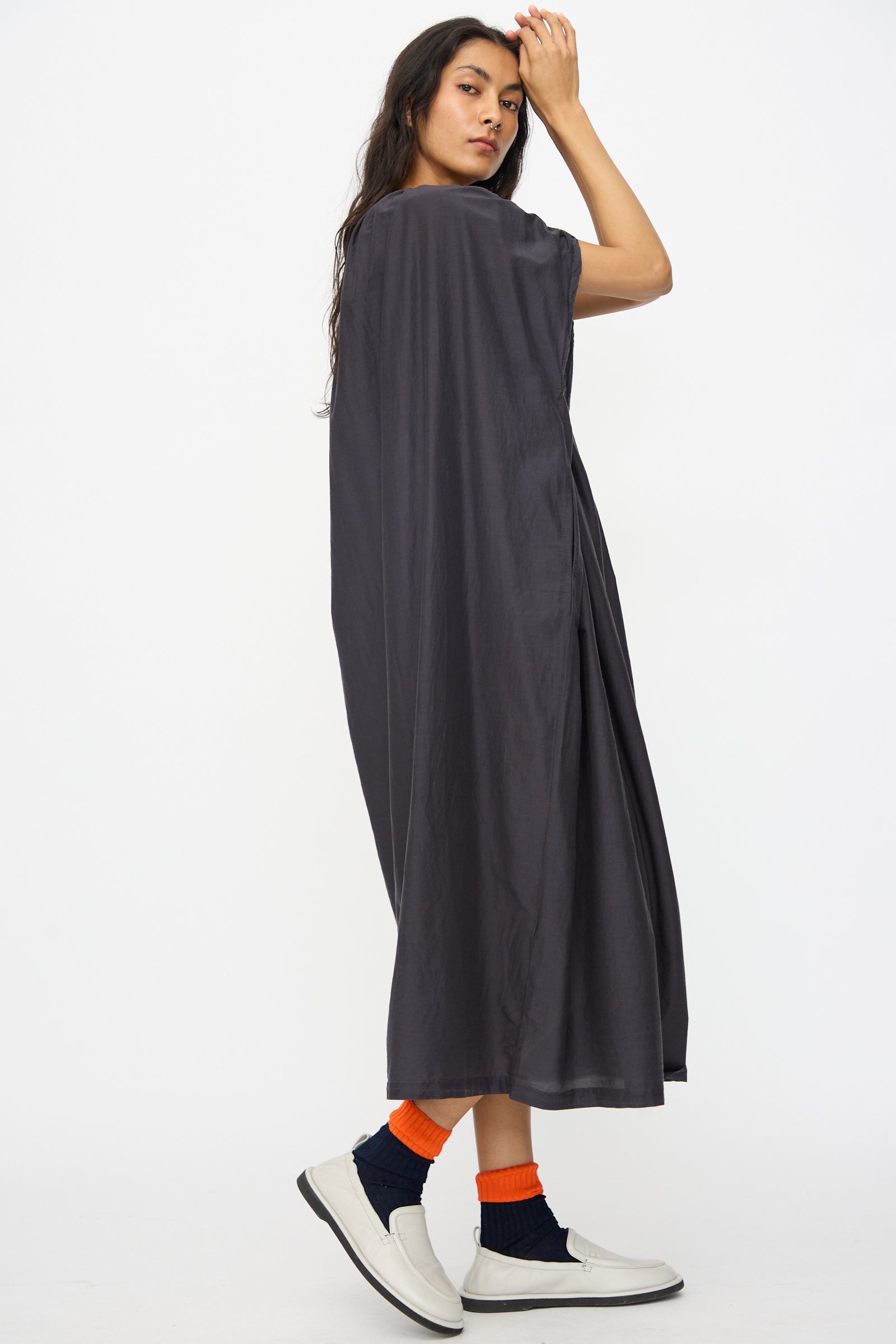 A woman stands sideways wearing the Black Crane Mont Blanc Dress in Faded Black, paired with navy socks with orange stripes and white slip-on shoes, set against a plain white background.