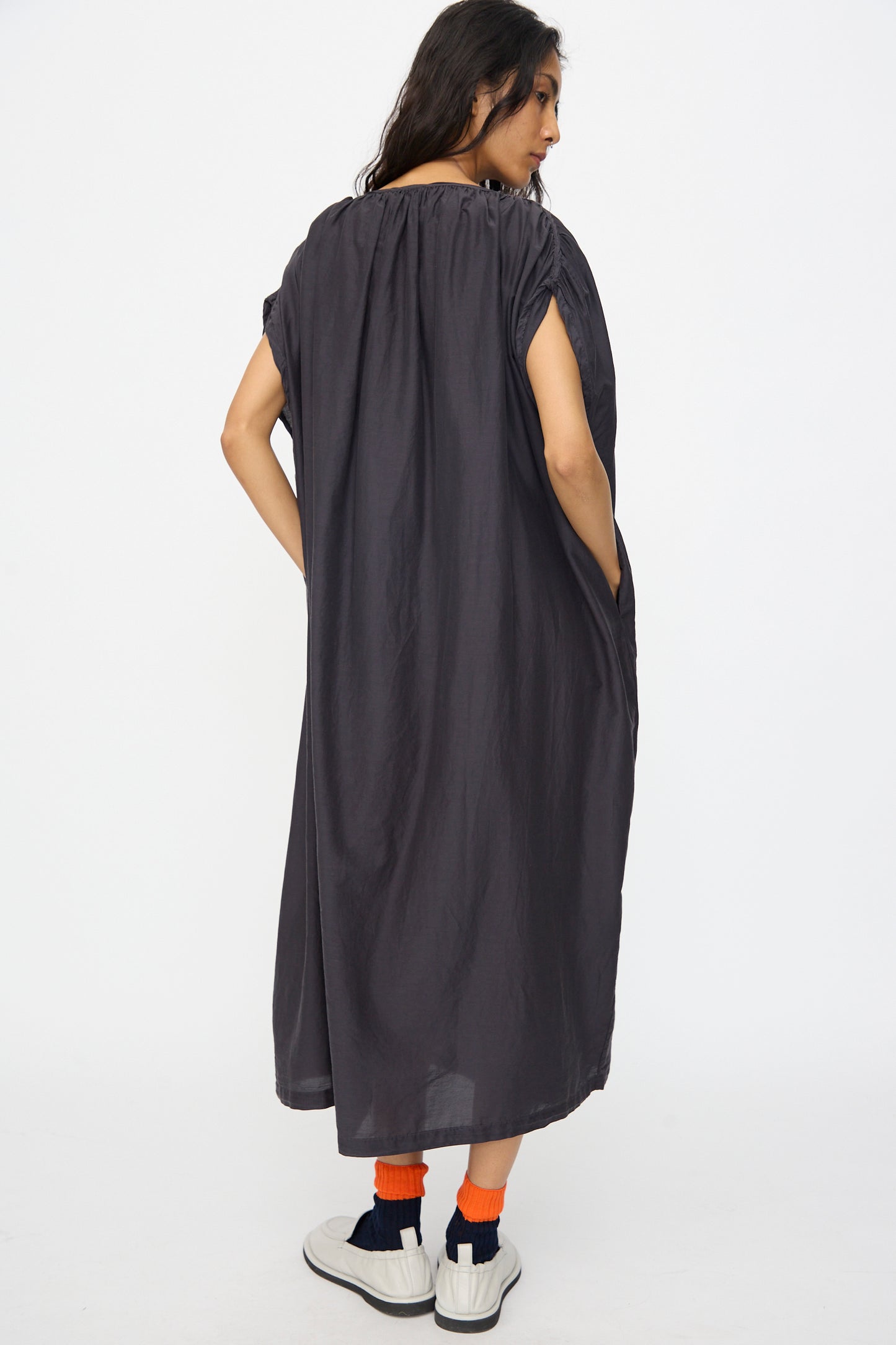 A person wearing the Black Crane Mont Blanc Dress in Faded Black, orange socks, and white sneakers stands facing away from the camera against a plain white background.