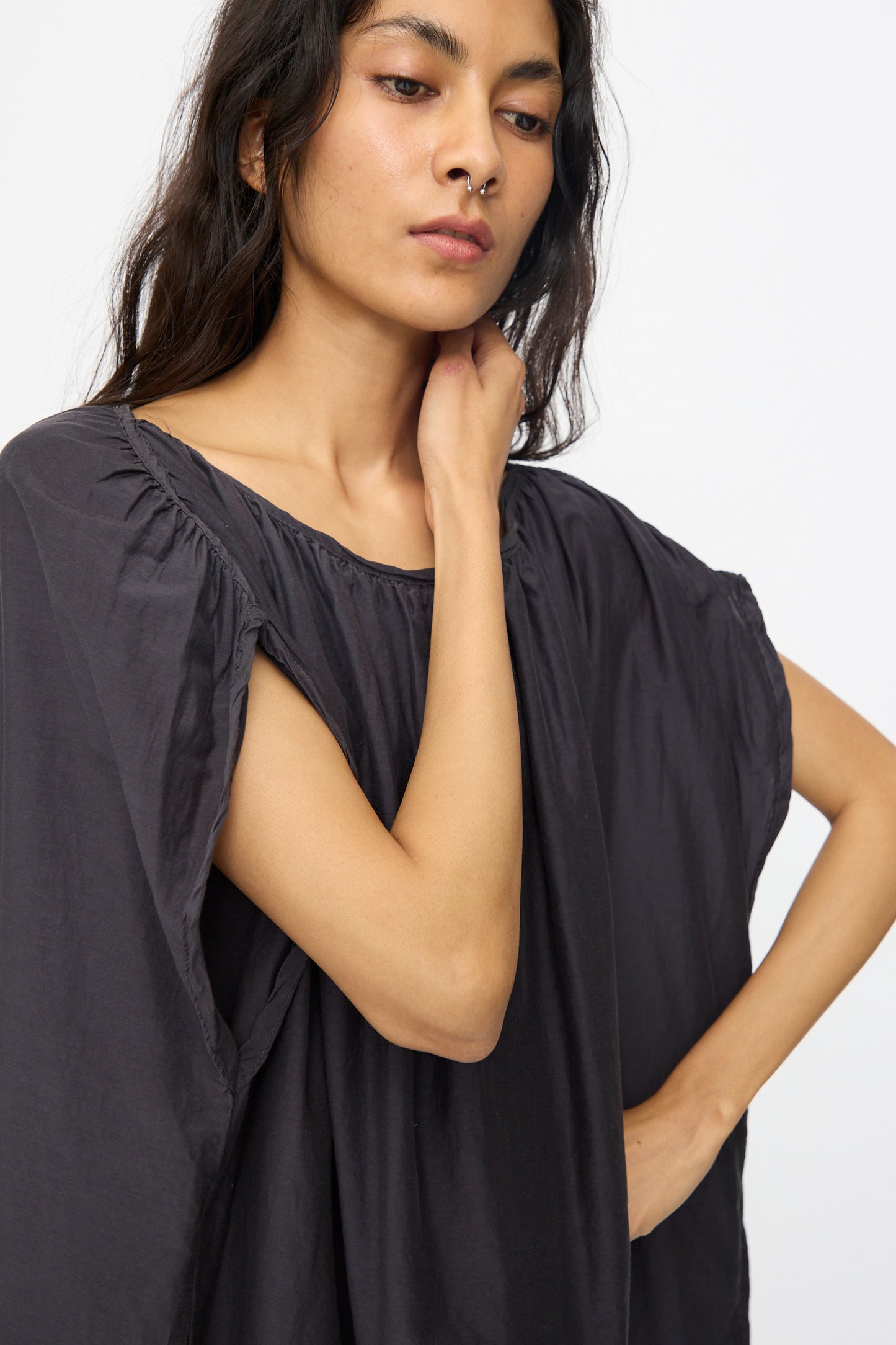 A woman with long dark hair, gazing downward and touching her neck, wears the Black Crane Mont Blanc Dress in faded black—a loose, short-sleeved piece with a wide silhouette.