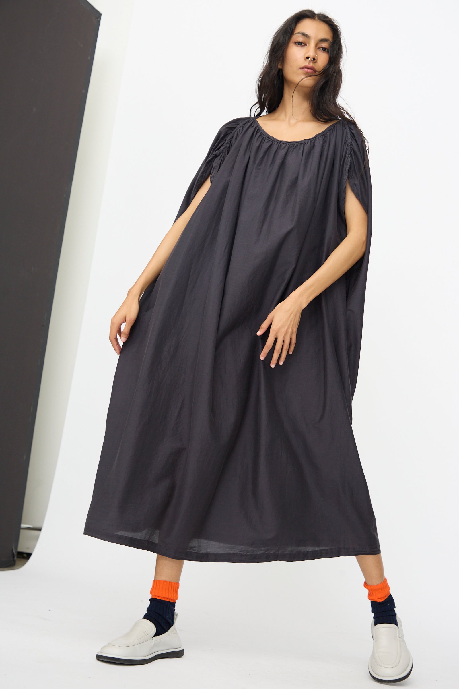 A person models the Black Crane Mont Blanc Dress in Faded Black, an oversized cotton silk blend with short sleeves, paired with orange socks, black leg warmers, and white shoes against a plain white background.