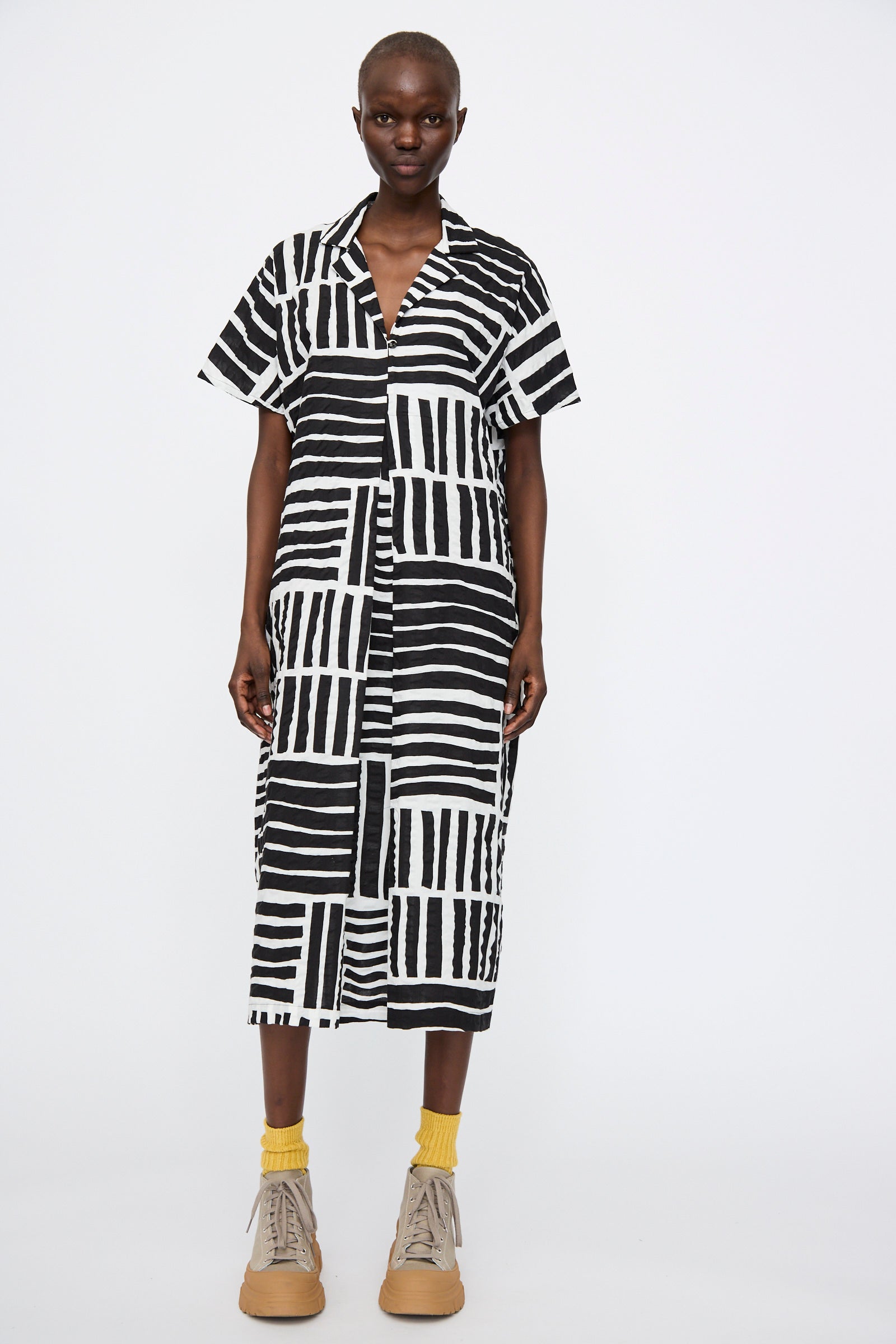 A person stands against a plain background wearing the Black Crane Narrow Kite Dress in Black Print, styled with yellow socks and beige sneakers.