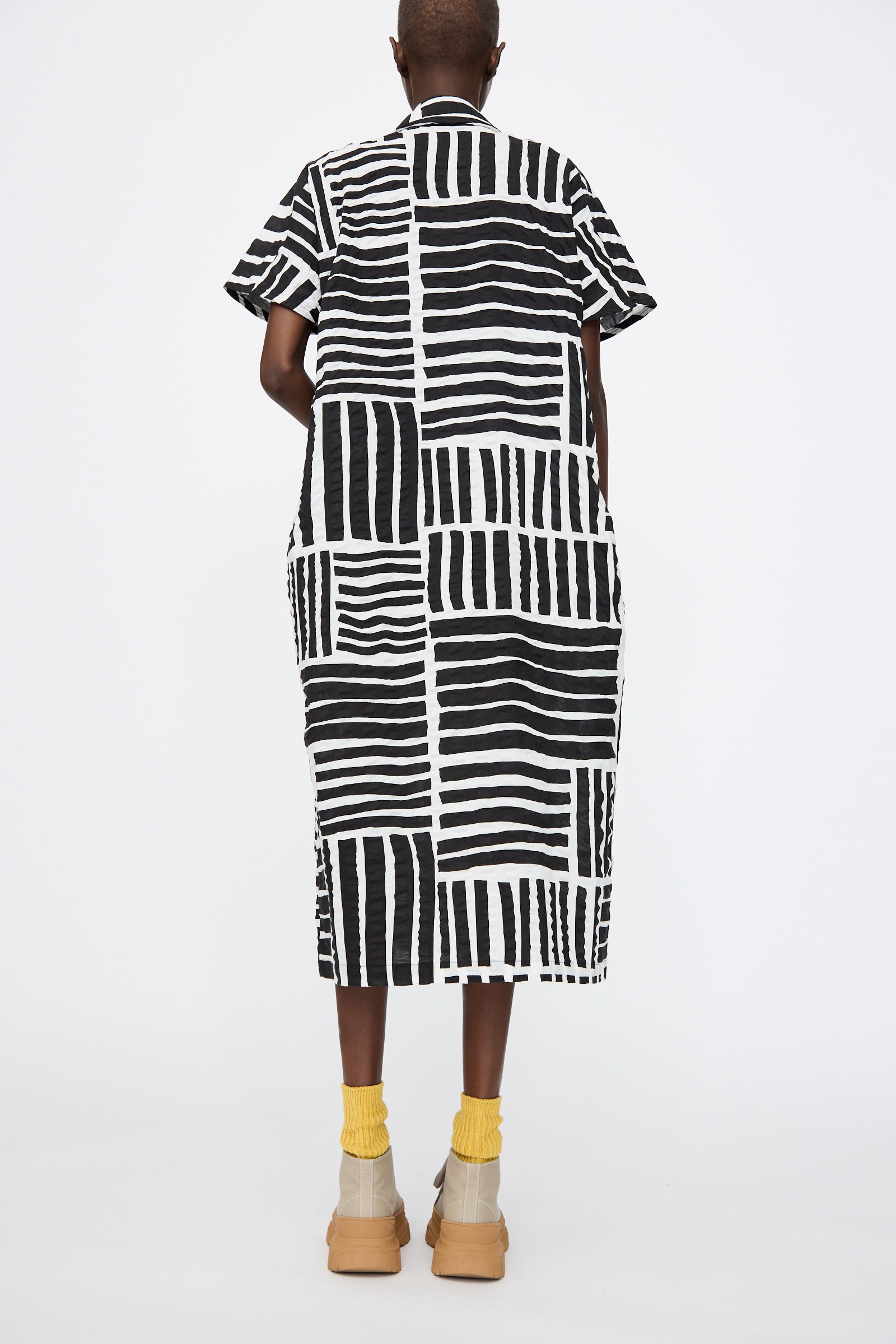 A person stands facing away against a white background, wearing the Black Crane Narrow Kite Dress in Black Print, styled with yellow socks and beige shoes.