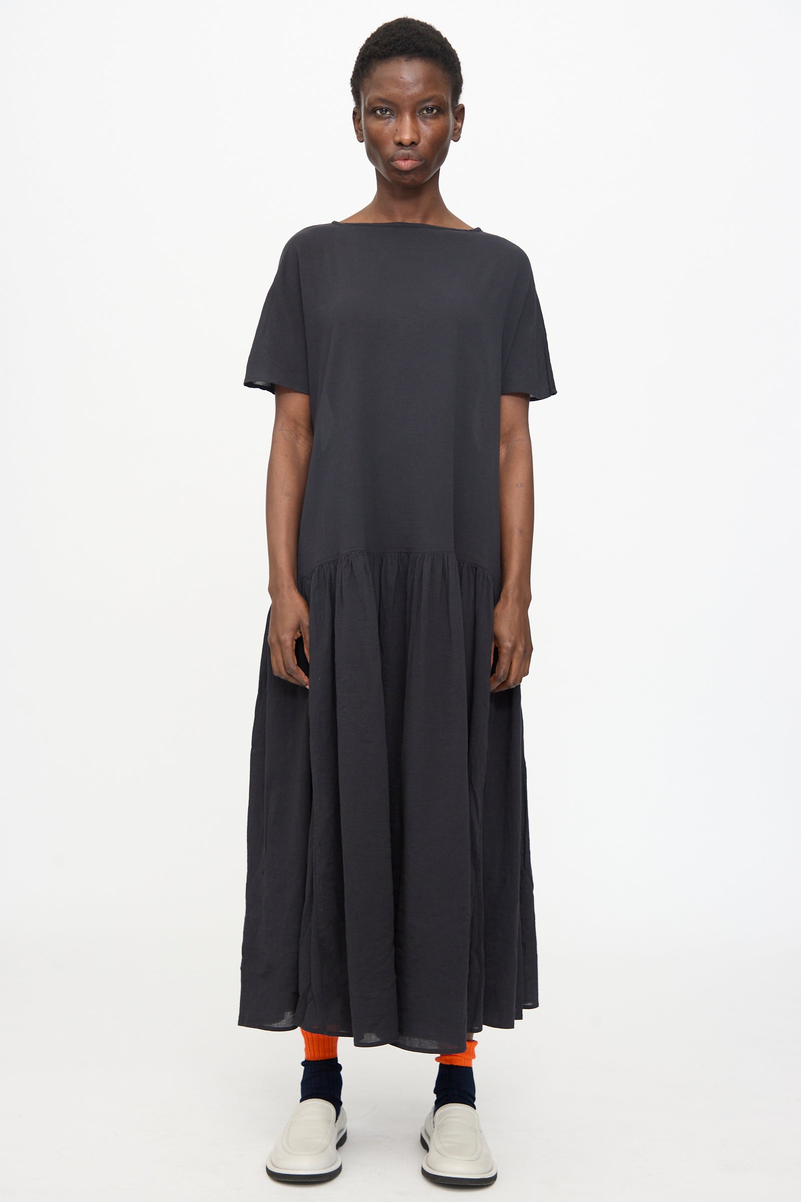 A person faces forward wearing the Black Crane New Easy Dress in Sumi Black—an oversized, short-sleeved cotton gauze dress—paired with orange socks and white slip-on shoes against a plain white background.