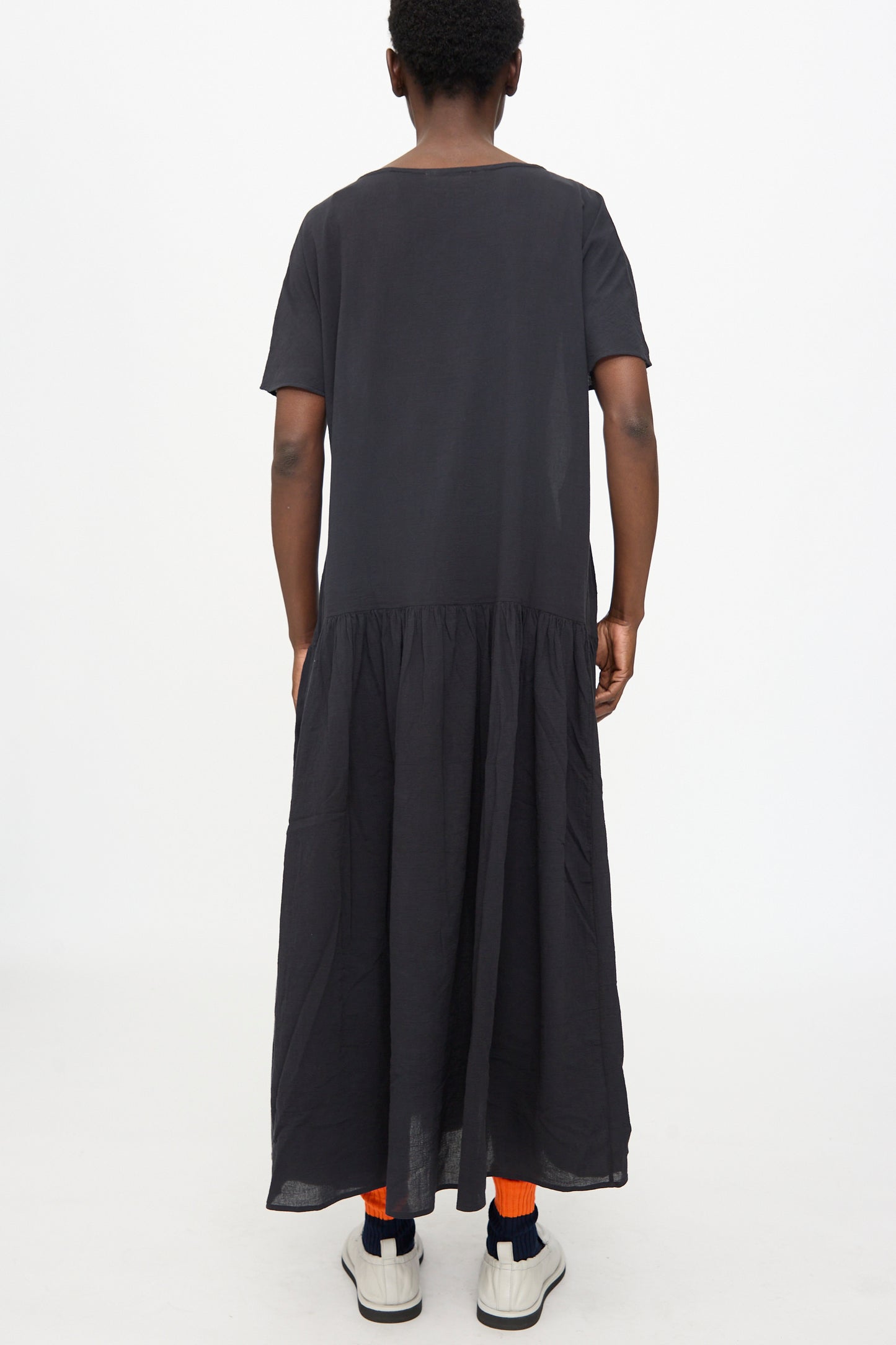 A person stands facing away against a plain white background, wearing the Black Crane New Easy Dress in Sumi Black, paired with orange socks and white sneakers.