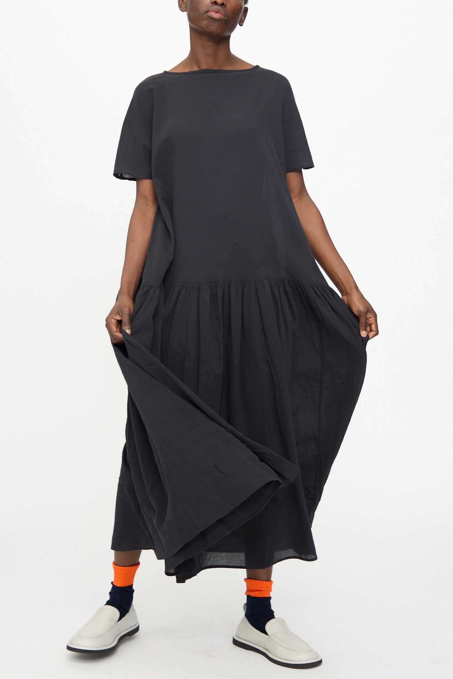 A person wears the Black Crane New Easy Dress in Sumi Black— a loose-fitting cotton gauze maxi dress with short sleeves—paired with orange socks and white slip-on shoes, standing against a plain white background.