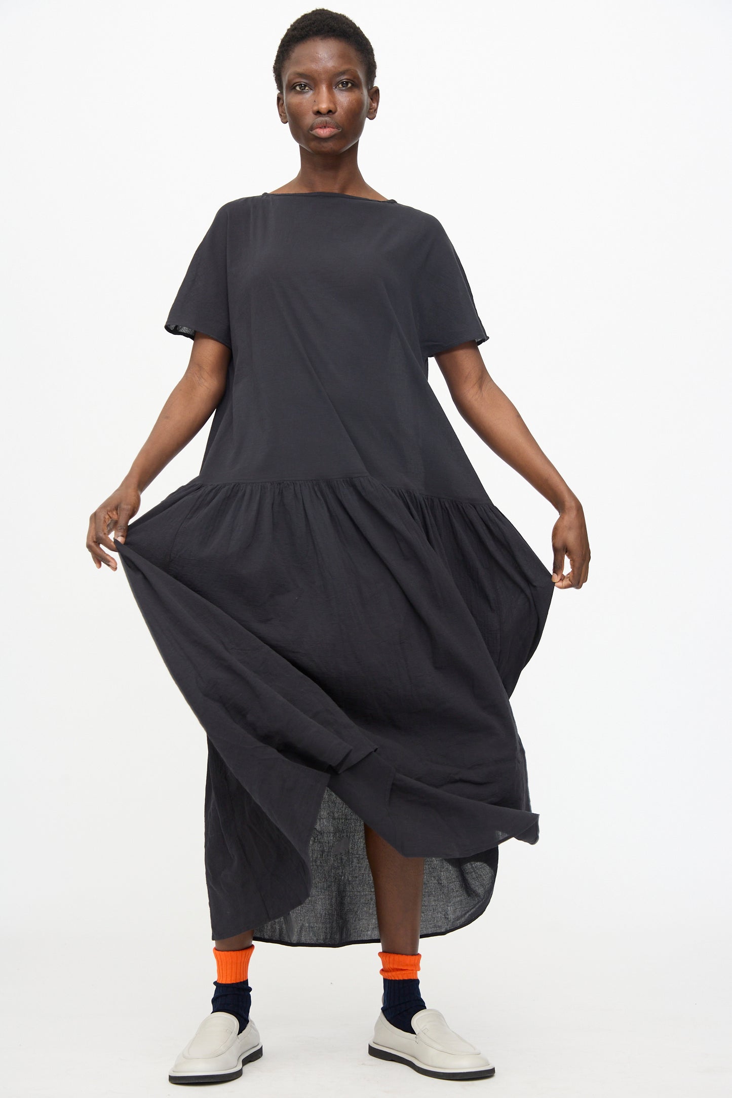 A person wearing the Black Crane New Easy Dress in Sumi Black, with orange socks and white slip-on shoes, stands against a plain white background holding out the skirt.