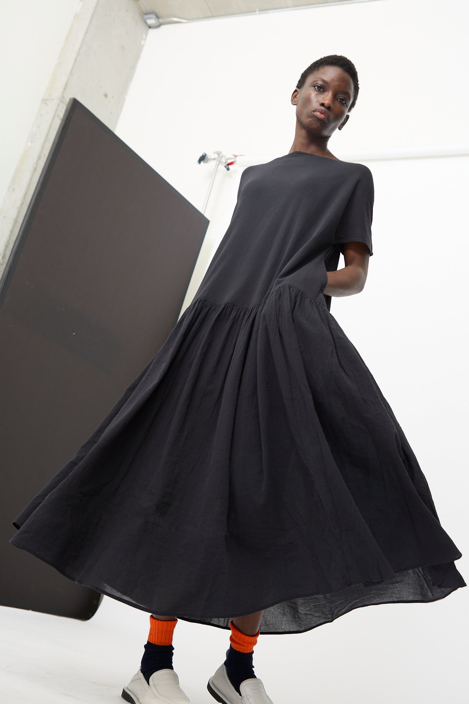 A person wears the Black Crane New Easy Dress in Sumi Black—an oversized, short-sleeved style with a flared skirt—paired with white socks and black shoes in a minimalist indoor setting.