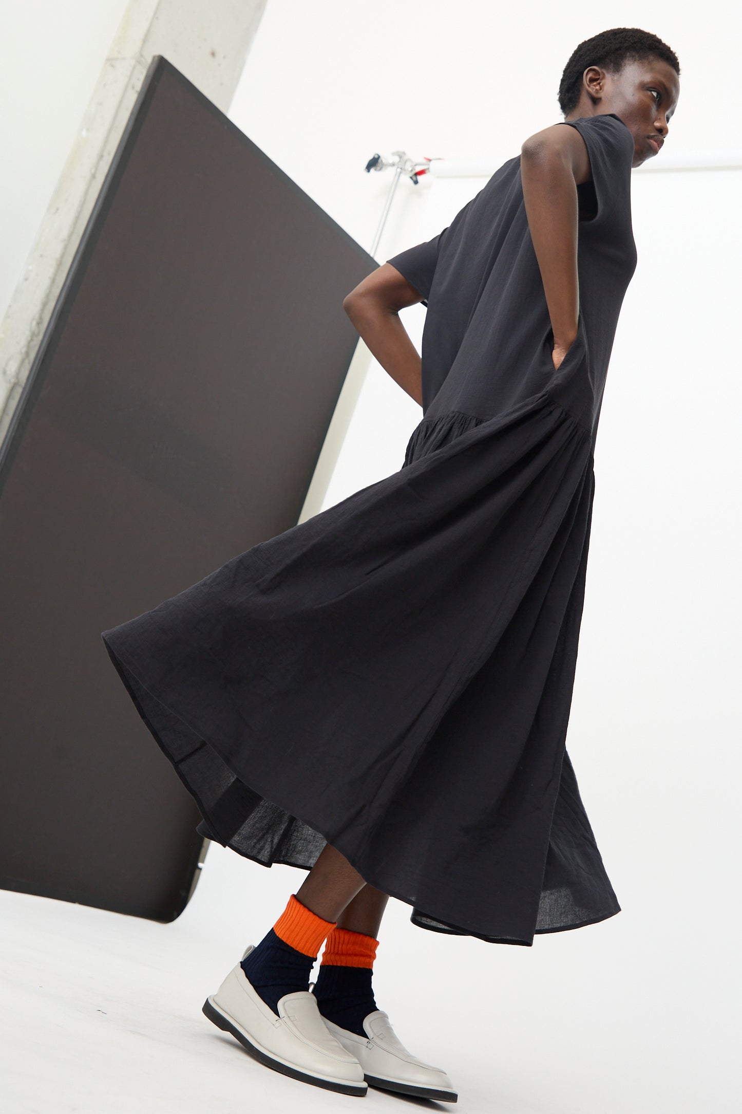 A person stands indoors with hands in pockets, wearing the Black Crane New Easy Dress in Sumi Black, orange and dark socks, and white slip-on shoes; white background with a dark panel.