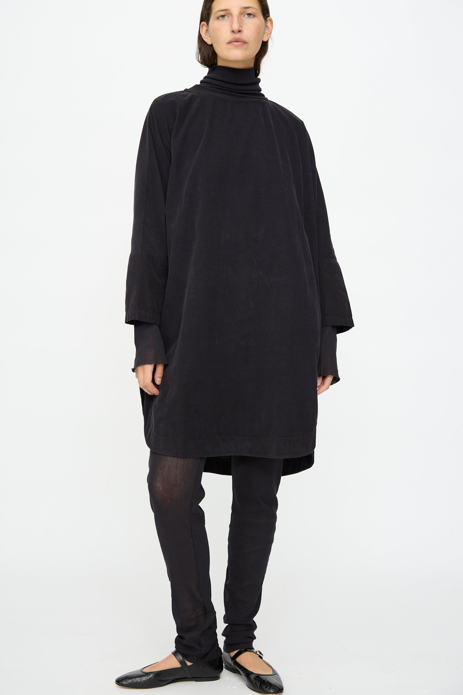 A person stands against a plain background wearing the Black Crane Painter Dress in Dark Navy, made in the USA, layered over a black turtleneck, black leggings, and black ballet flats.