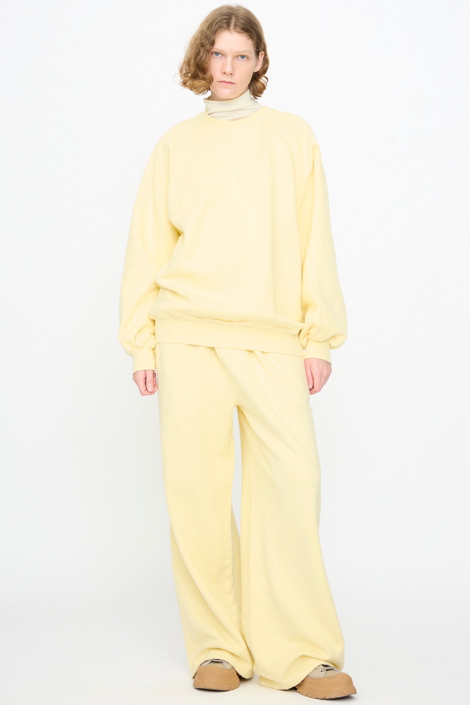 A person stands against a white background, wearing the Black Crane Raglan Sweatshirt in Custard—a loose pullover crafted from an organic cotton blend—paired with matching wide-leg pants and tan shoes.