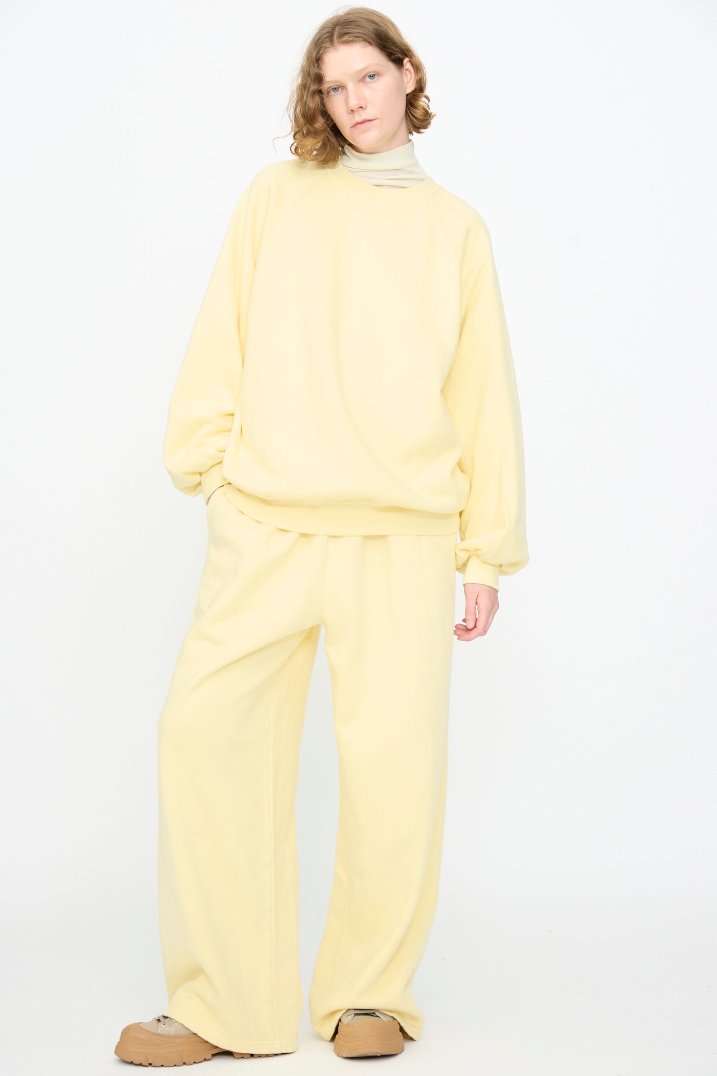 A person stands against a white background, wearing Black Crane’s Raglan Sweatshirt in Custard—an oversized pullover crafted from organic cotton—paired with matching wide-leg pants, a beige turtleneck, and tan shoes.