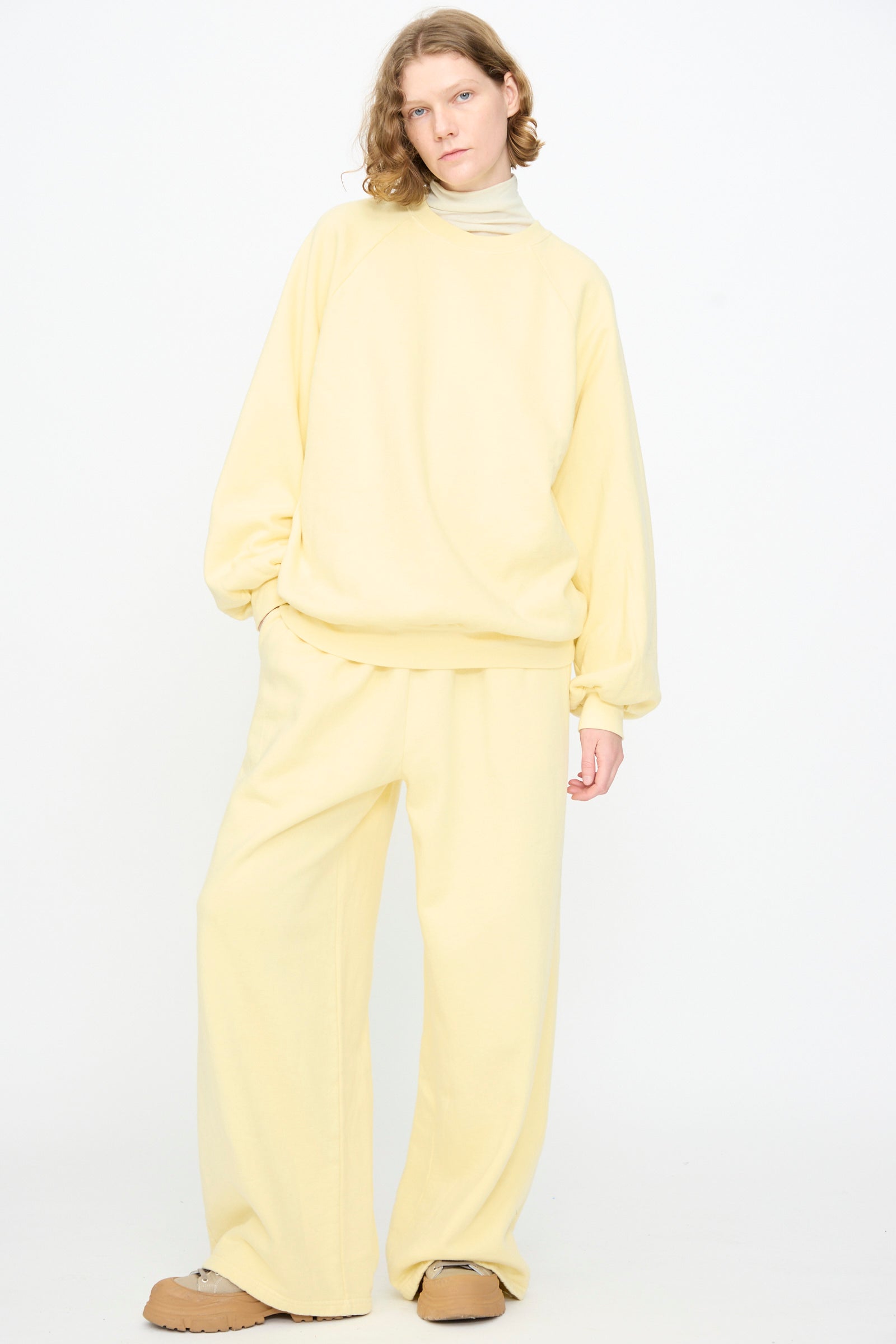 A person stands against a white background, wearing Black Crane’s Raglan Sweatshirt in Custard—an oversized pullover crafted from organic cotton—paired with matching wide-leg pants, a beige turtleneck, and tan shoes.