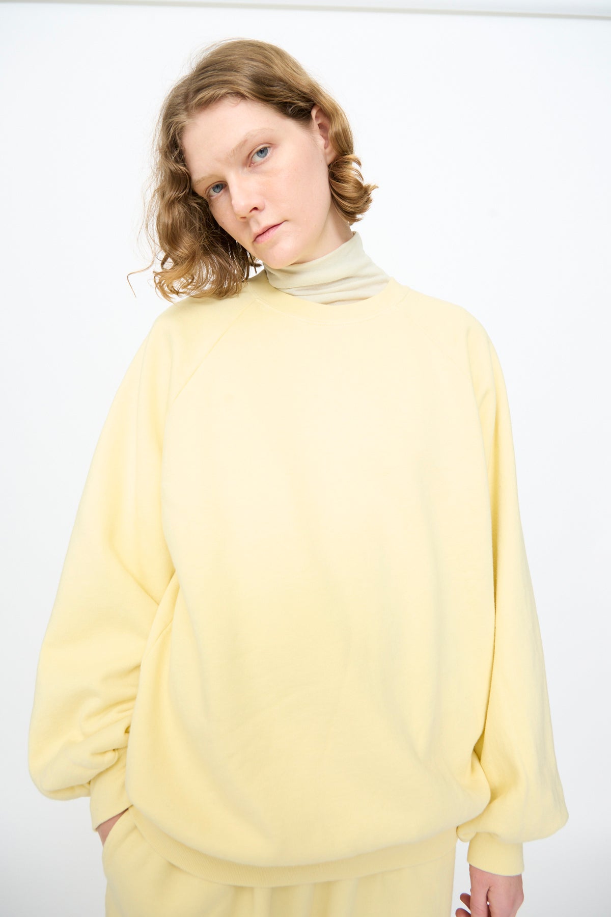 A person with light brown, wavy hair wears the Black Crane Raglan Sweatshirt in Custard—an oversized pullover crafted from organic cotton—layered over a light turtleneck, standing against a plain white background.