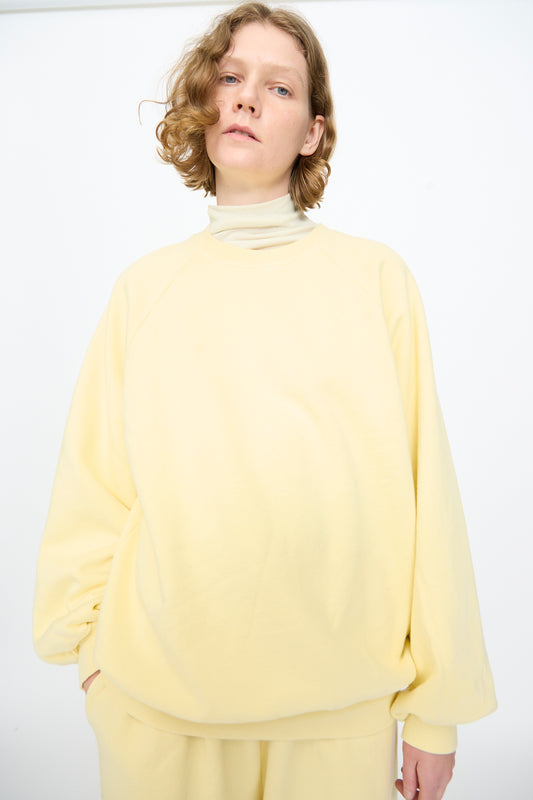 A person with short wavy hair wears the Black Crane Raglan Sweatshirt in Custard— a loose-fitting, pale yellow pullover crafted from soft organic cotton— paired with matching pants, standing against a plain white background.