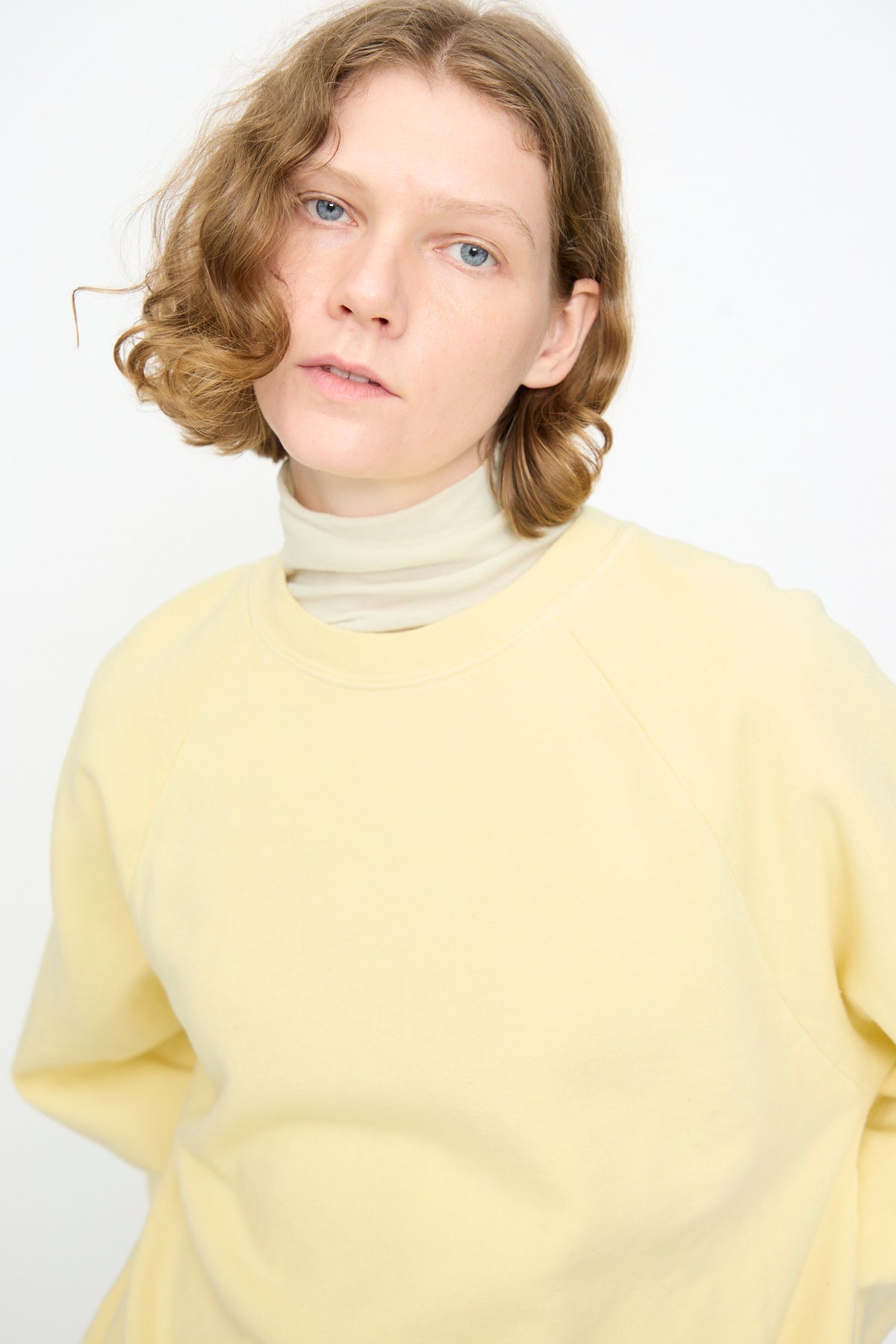 A person with short, wavy light brown hair wears the Black Crane Raglan Sweatshirt in Custard—an organic cotton pullover—layered over a beige turtleneck, standing against a plain white background and looking directly at the camera.