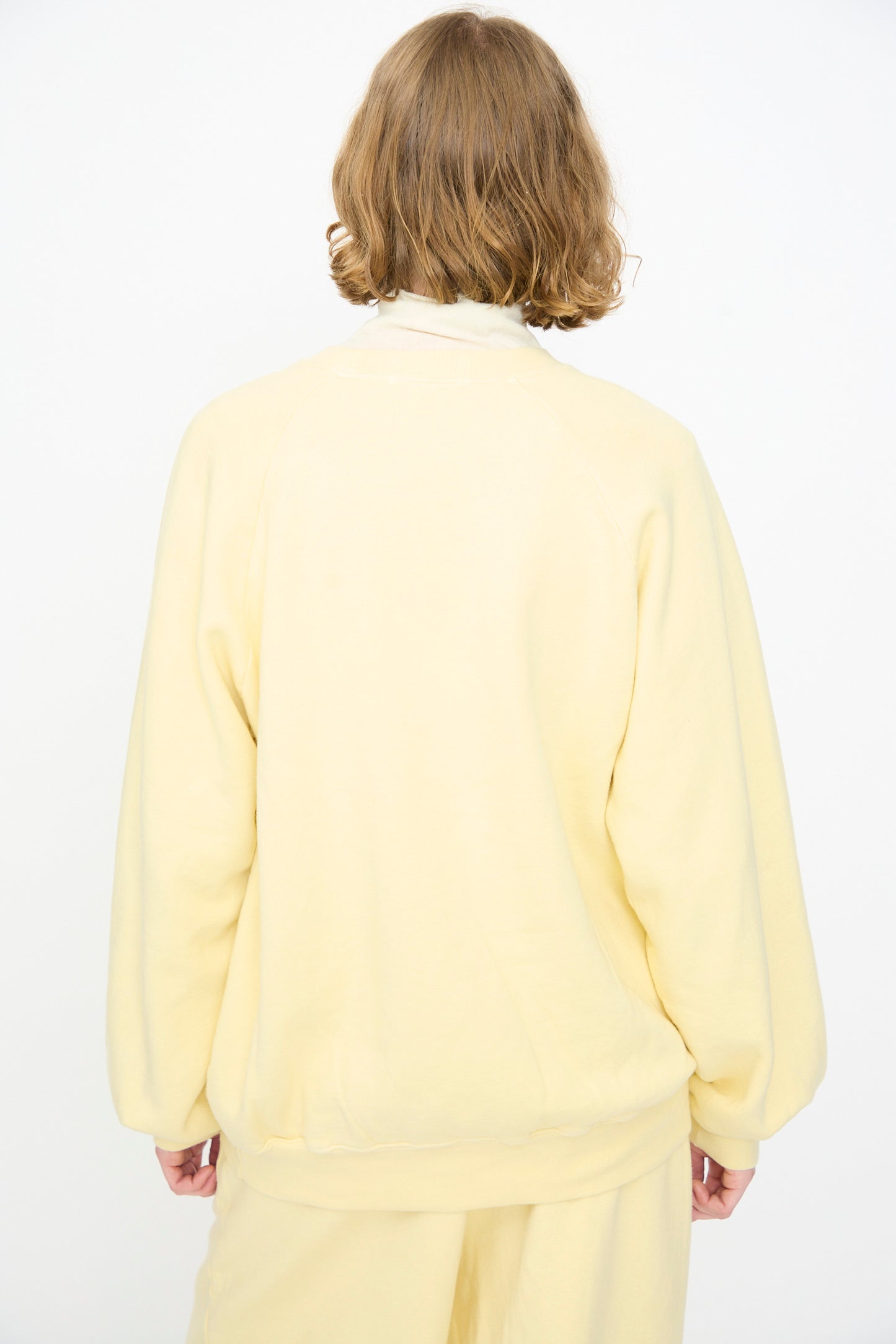 A person with short, wavy brown hair is shown from the back against a plain white background, wearing the Black Crane Raglan Sweatshirt in Custard paired with matching pants.