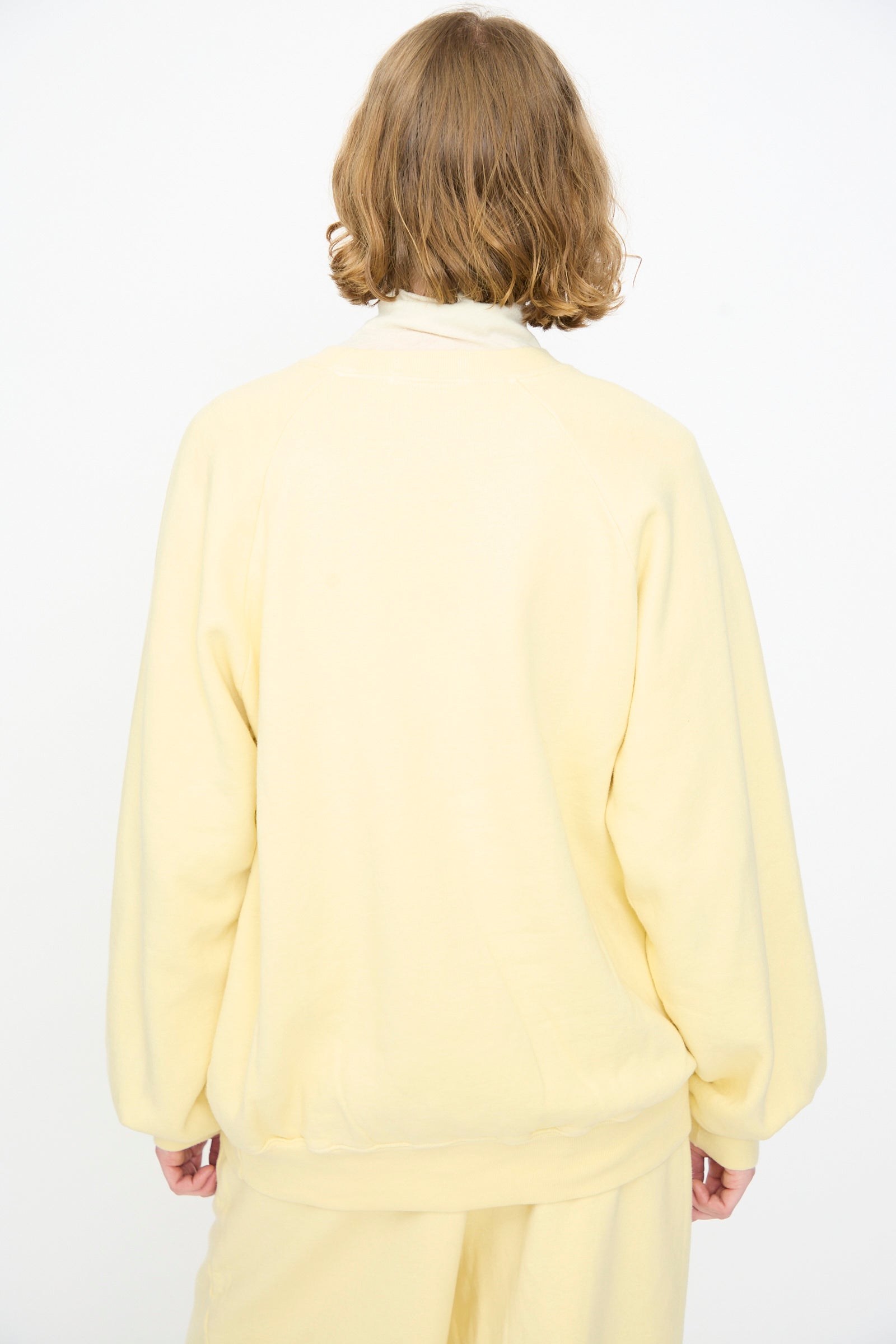 A person with short, wavy brown hair is shown from the back against a plain white background, wearing the Black Crane Raglan Sweatshirt in Custard paired with matching pants.