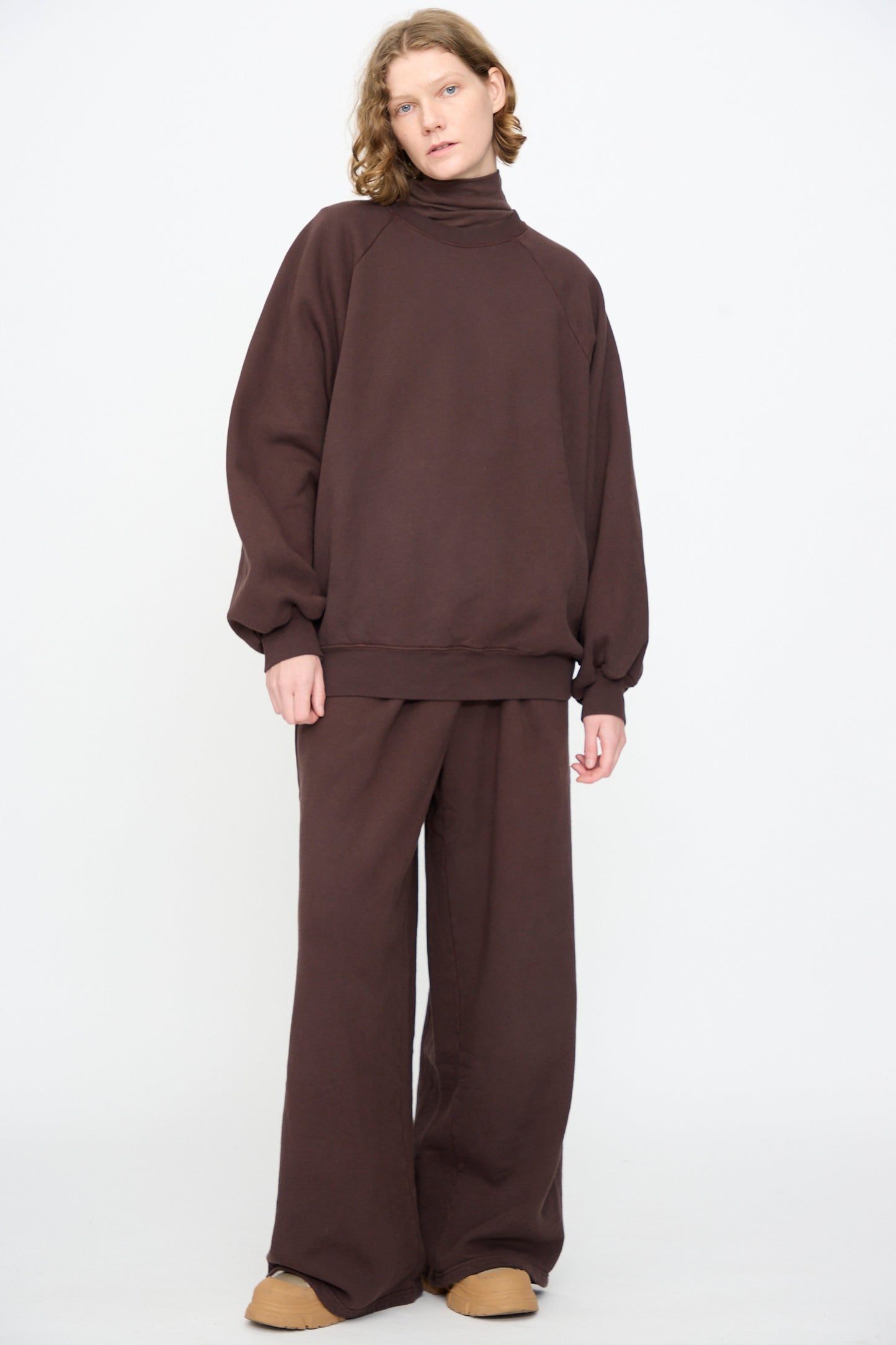 A person wearing the Black Crane Raglan Sweatshirt in Dark Cacao—an oversized pullover crafted from a soft cotton-cashmere blend—with coordinating wide-leg pants, standing against a plain white background.
