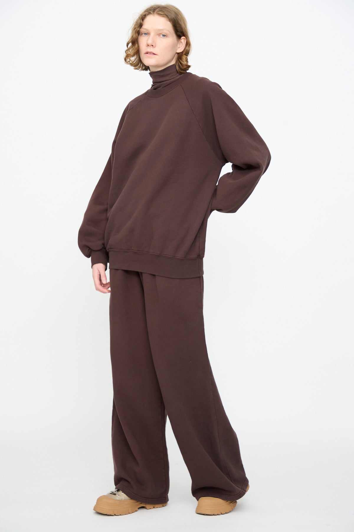 A person stands against a white background, wearing the Black Crane Raglan Sweatshirt in Dark Cacao—a loose turtleneck crafted from a soft cotton cashmere blend—paired with matching wide-leg pants and tan slip-on shoes.