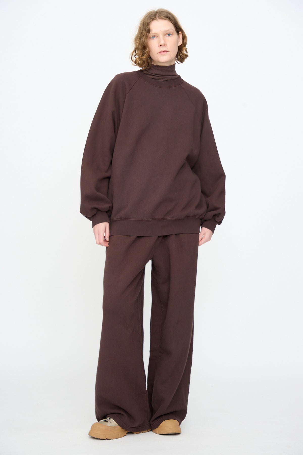 Person standing against a plain white background, wearing the Black Crane Raglan Sweatshirt in Dark Cacao—a loose-fitting pullover crafted from organic cotton—paired with matching wide-leg pants and tan shoes.
