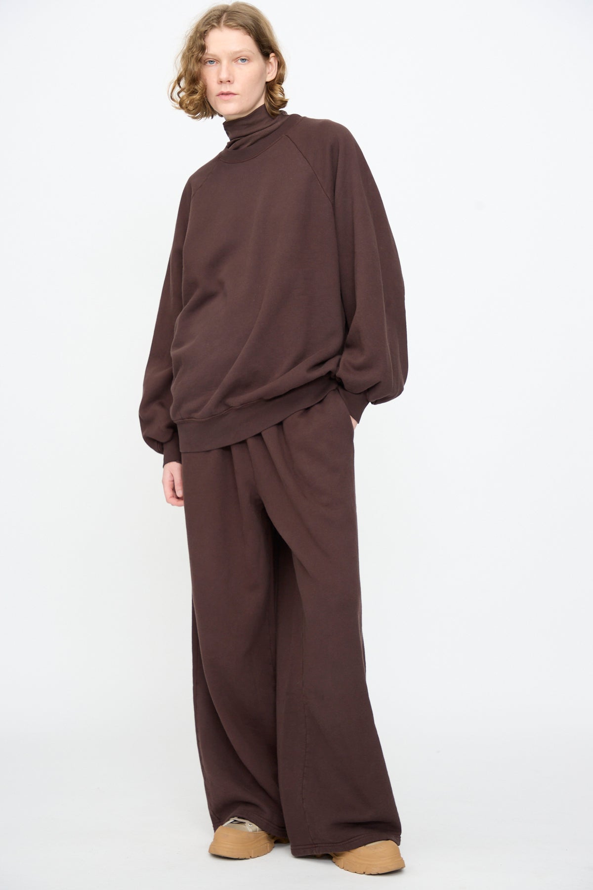 A person wearing the Black Crane Raglan Sweatshirt in Dark Cacao and wide-leg brown pants, standing against a plain white background.