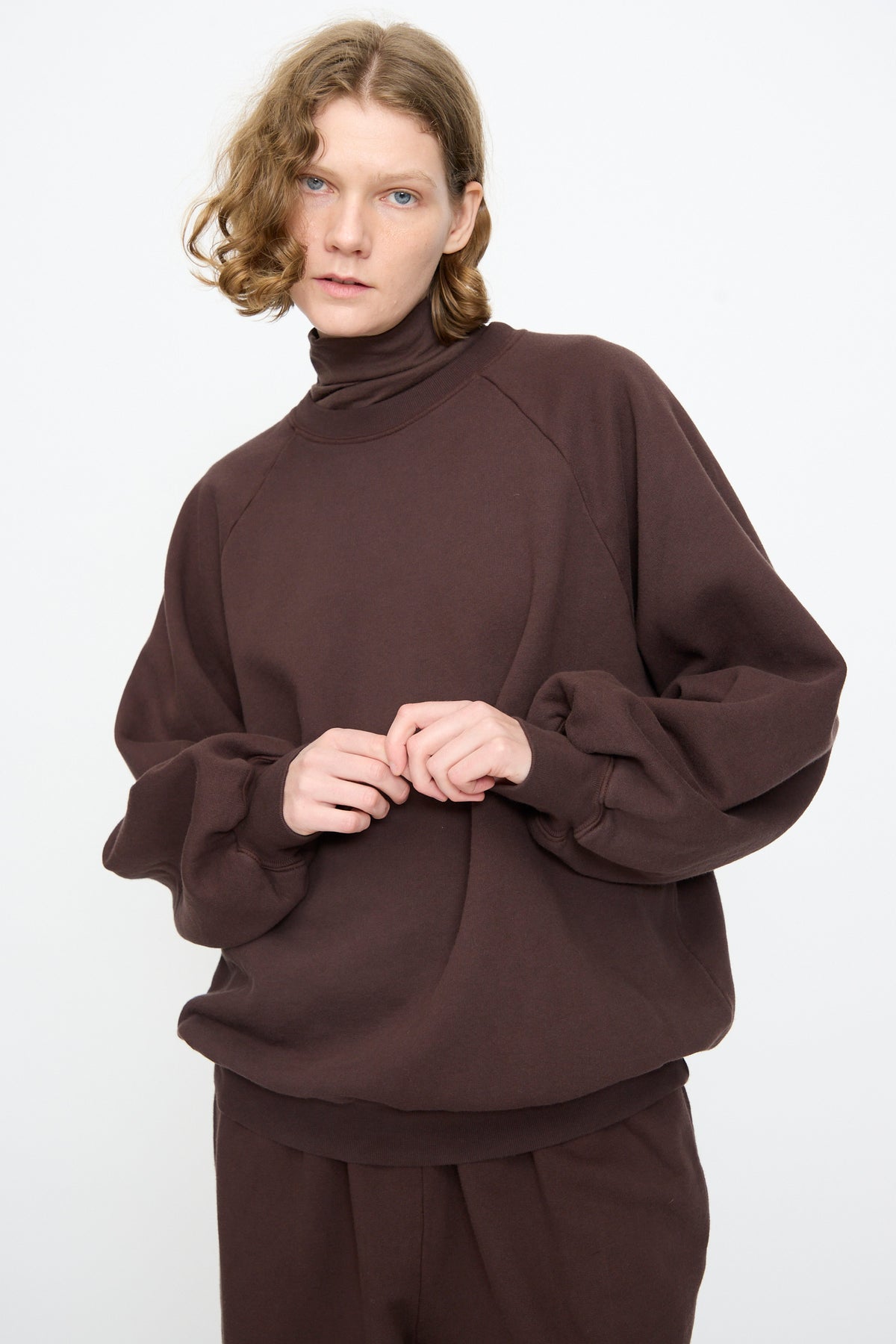 A person with short, wavy blond hair wears the Black Crane Raglan Sweatshirt in Dark Cacao with matching pants, standing against a plain white background.