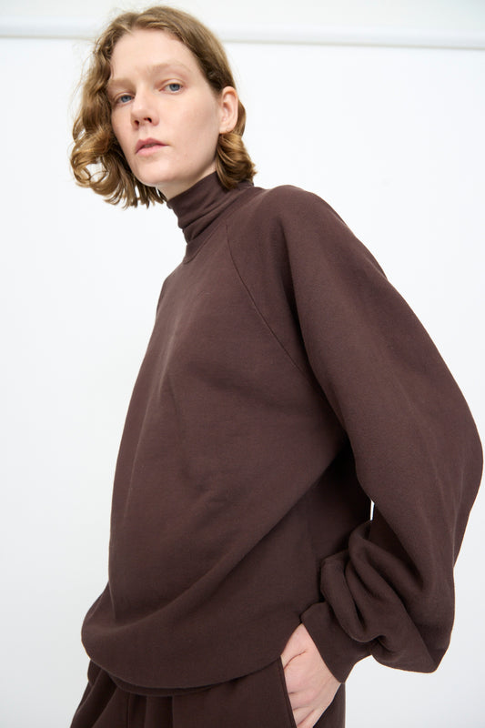 A person with short, wavy, light brown hair wears the Black Crane Raglan Sweatshirt in Dark Cacao—a soft cotton cashmere blend pullover—paired with matching pants, standing against a plain white background.