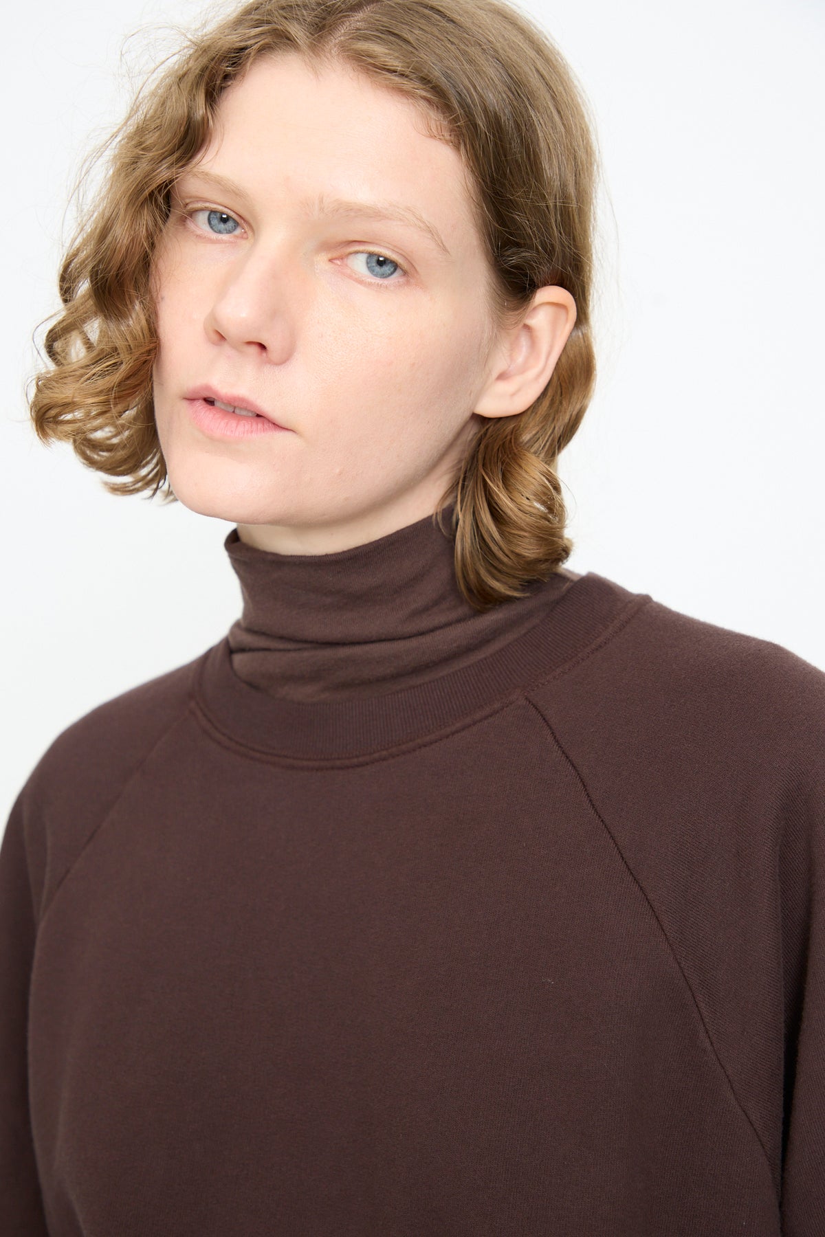 Person with wavy, shoulder-length blonde hair wearing the Black Crane Raglan Sweatshirt in Dark Cacao, looking slightly past the camera against a plain white background.