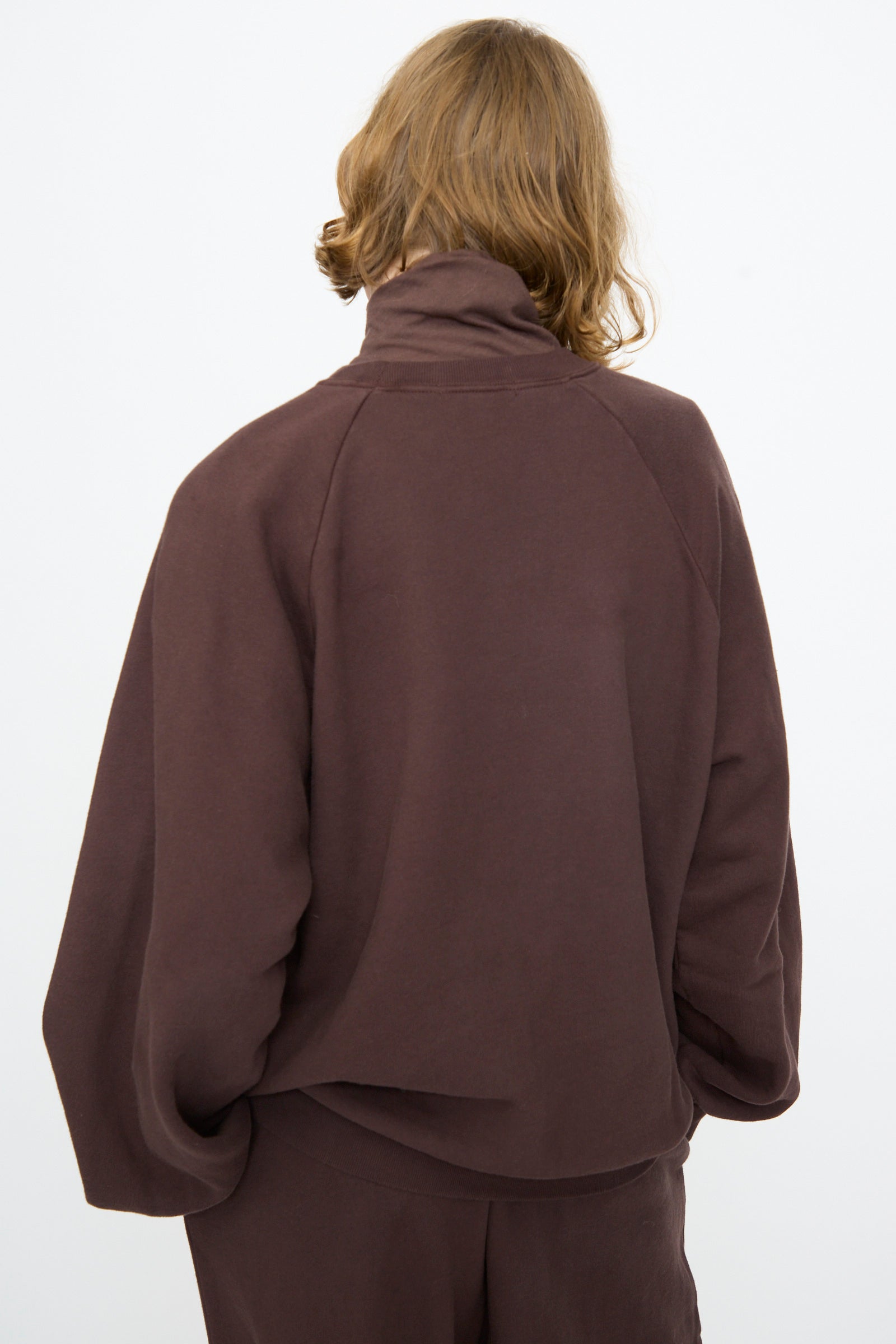 A person with shoulder-length light brown hair is seen from behind, wearing the Black Crane Raglan Sweatshirt in Dark Cacao paired with matching pants, standing against a plain white background.