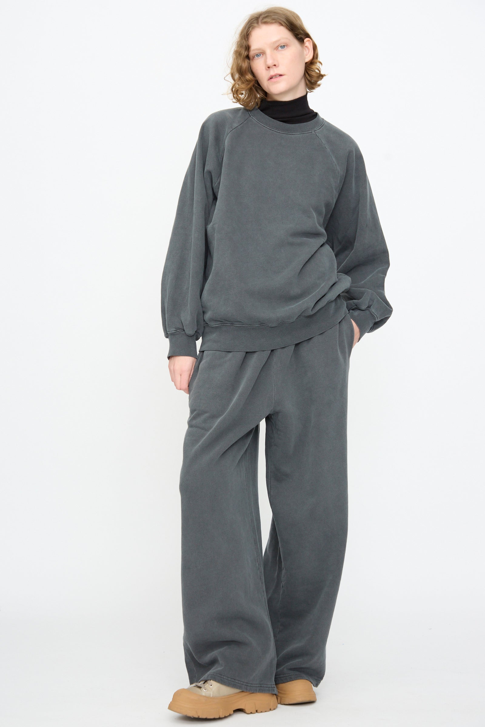 A person stands against a white background wearing the Black Crane Raglan Sweatshirt in Indigo, paired with matching wide-leg pants, a black turtleneck, and tan shoes.