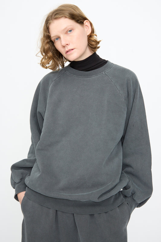 A person with shoulder-length blond hair wears the Black Crane Raglan Sweatshirt in Indigo, styled with matching pants and a black turtleneck, standing against a plain white background.