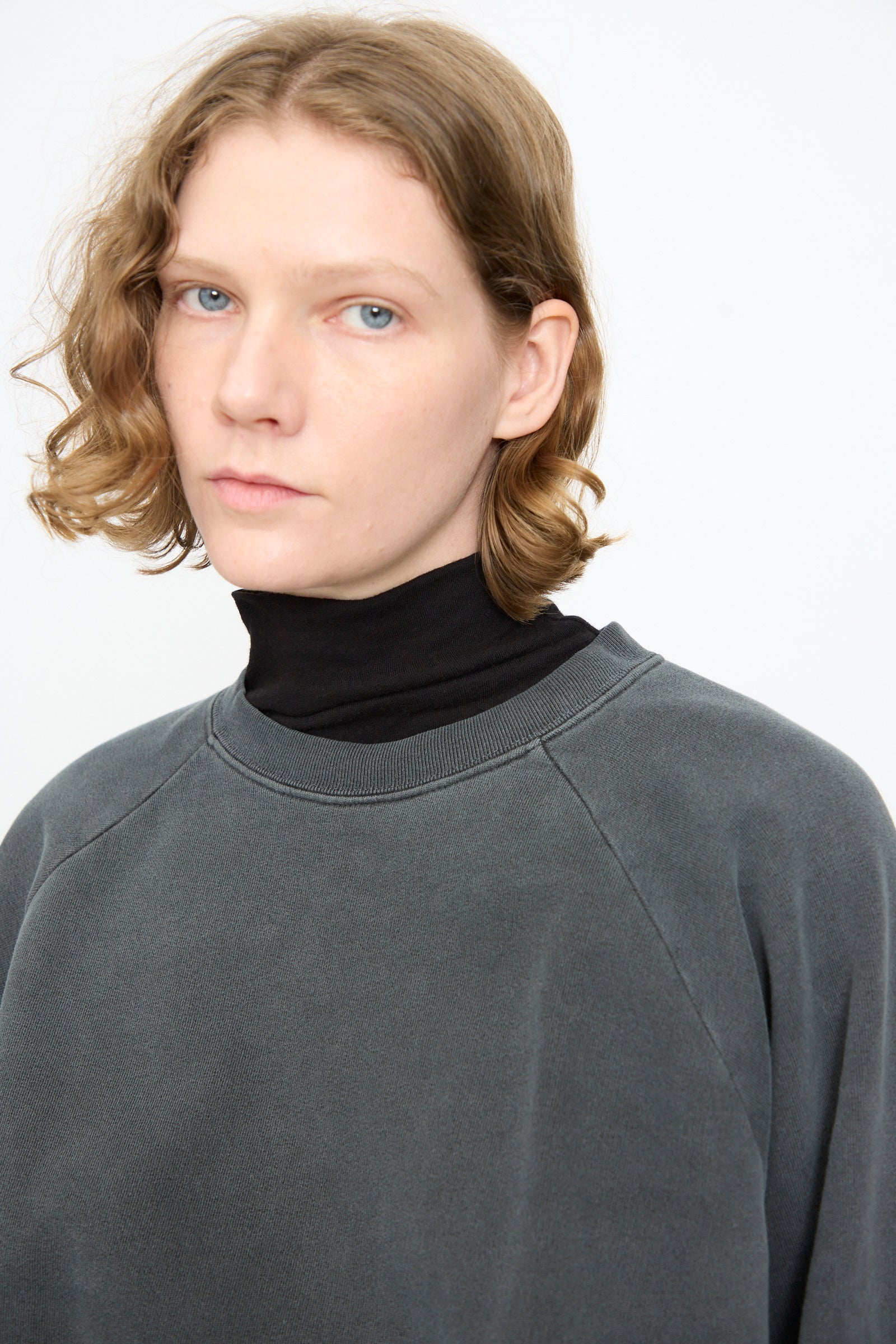 A person with short, wavy light brown hair faces the camera with a neutral expression against a plain light background, wearing the Black Crane Raglan Sweatshirt in Indigo over a black turtleneck.