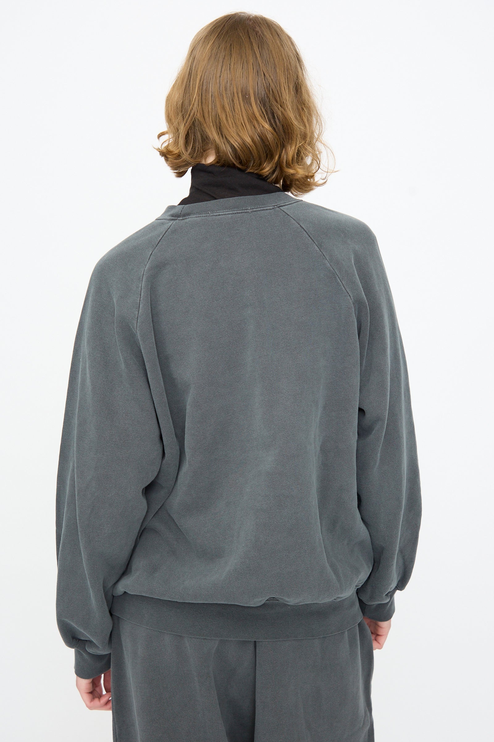 A person with shoulder-length light brown hair, wearing the Black Crane Raglan Sweatshirt in Indigo and matching pants, is standing with their back to the camera against a plain white background.