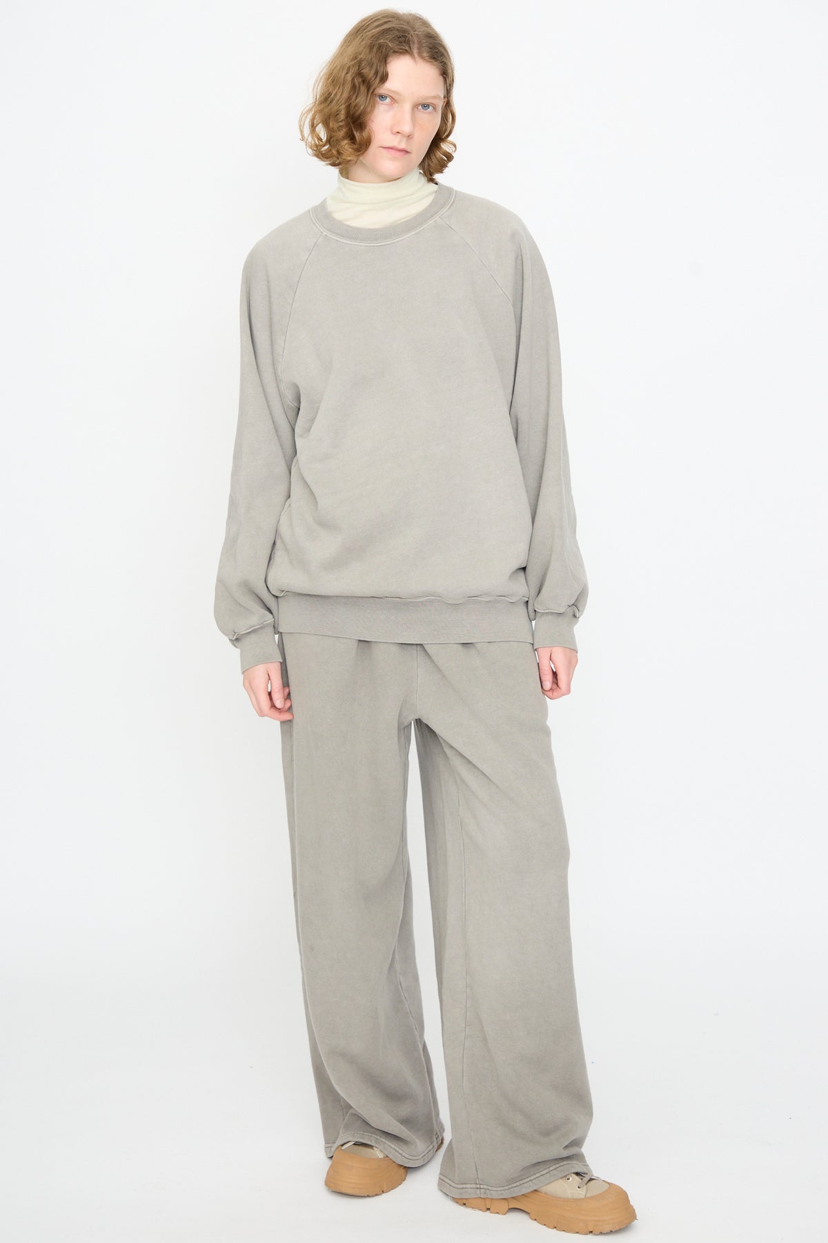 A person stands against a plain white background, wearing the Black Crane Raglan Sweatshirt in Stone — a loose-fitting, light gray cotton cashmere blend pullover — paired with matching wide-leg pants and tan shoes.