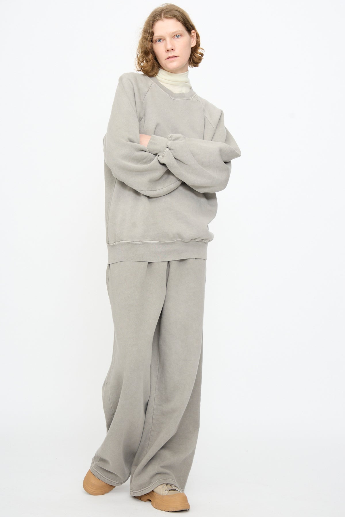 A person standing with arms crossed, wearing the Black Crane Raglan Sweatshirt in Stone—an oversized light gray organic cotton pullover—paired with matching pants, beige shoes, and a white turtleneck against a plain white background.