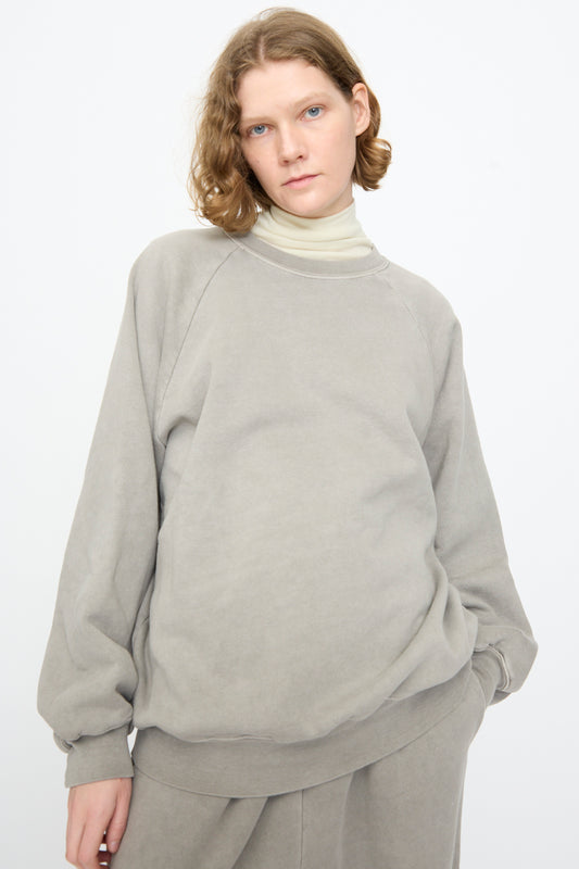 A person with shoulder-length light brown hair wears the Black Crane Raglan Sweatshirt in Stone, crafted from an organic cotton cashmere blend, layered over a beige turtleneck and paired with matching pants, standing against a plain light background.